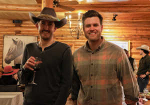 Two men smiling at the camera, one is wearing a cowboy hat and holding a glass of wine