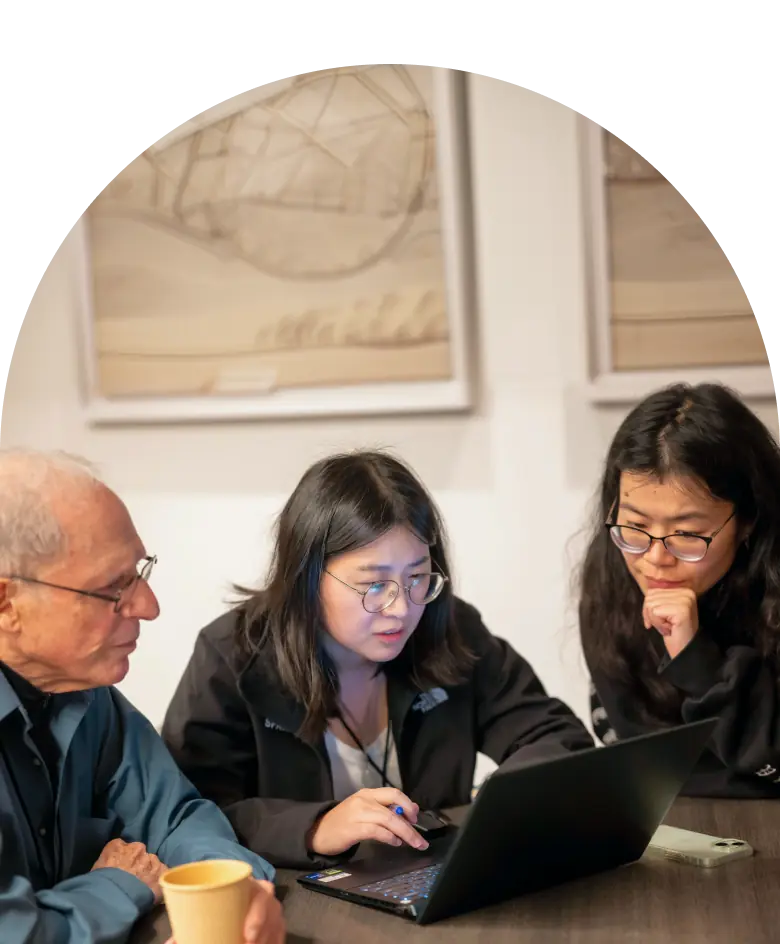 Three colleagues at a meeting table focused on a laptop, one woman pointing at the screen while the others look on.