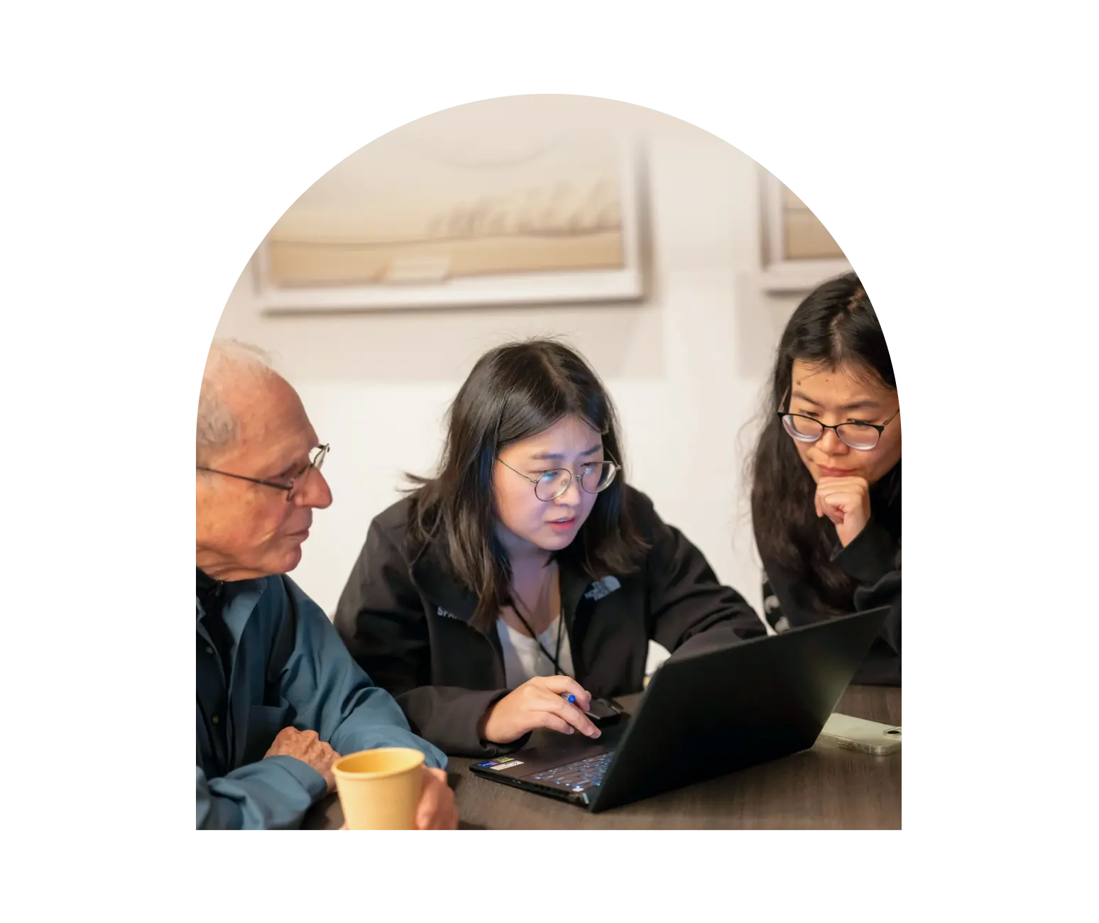 Three colleagues at a meeting table focused on a laptop, one woman pointing at the screen while the others look on.