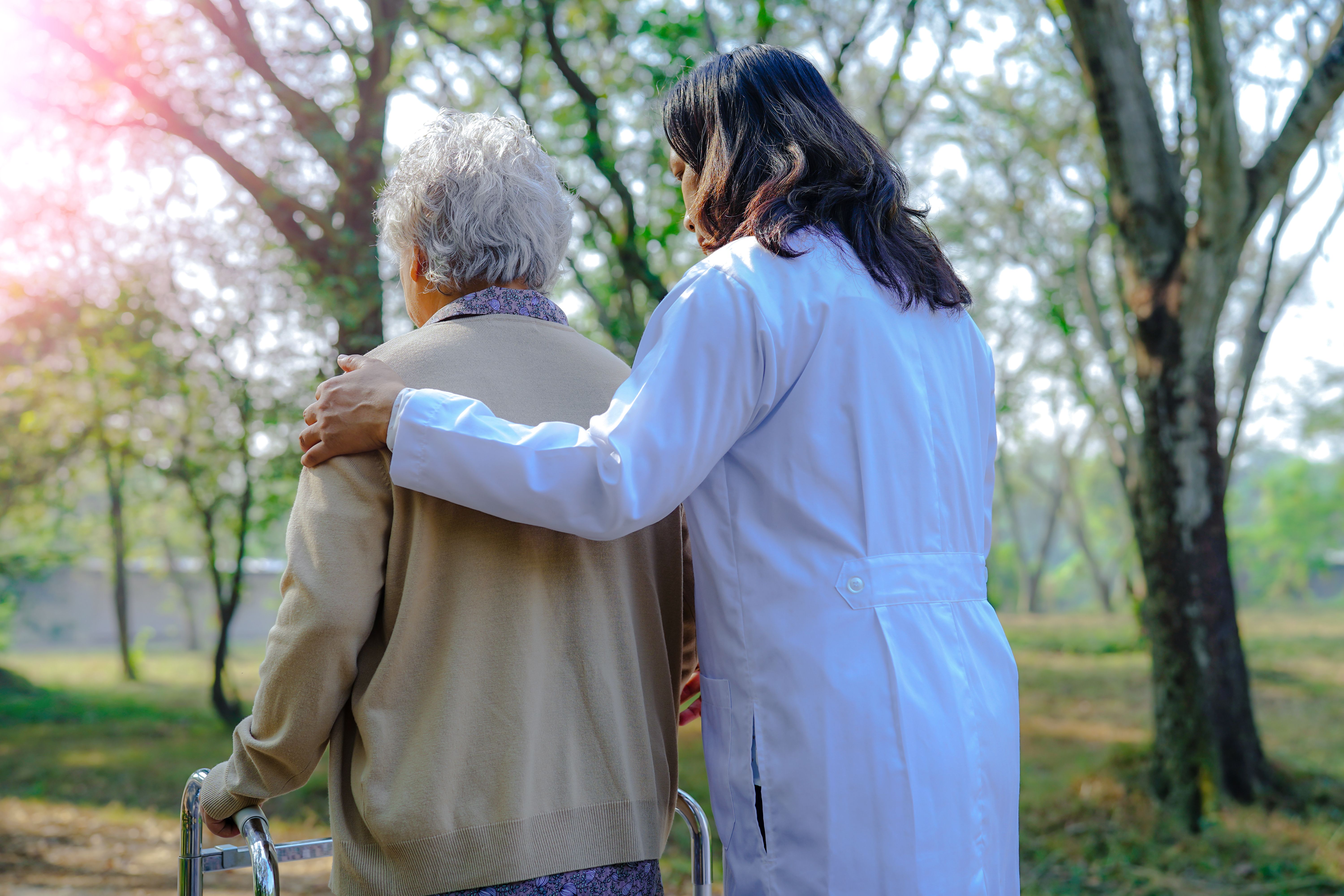 A doctor helping an elderly woman walk, viewed from behind.