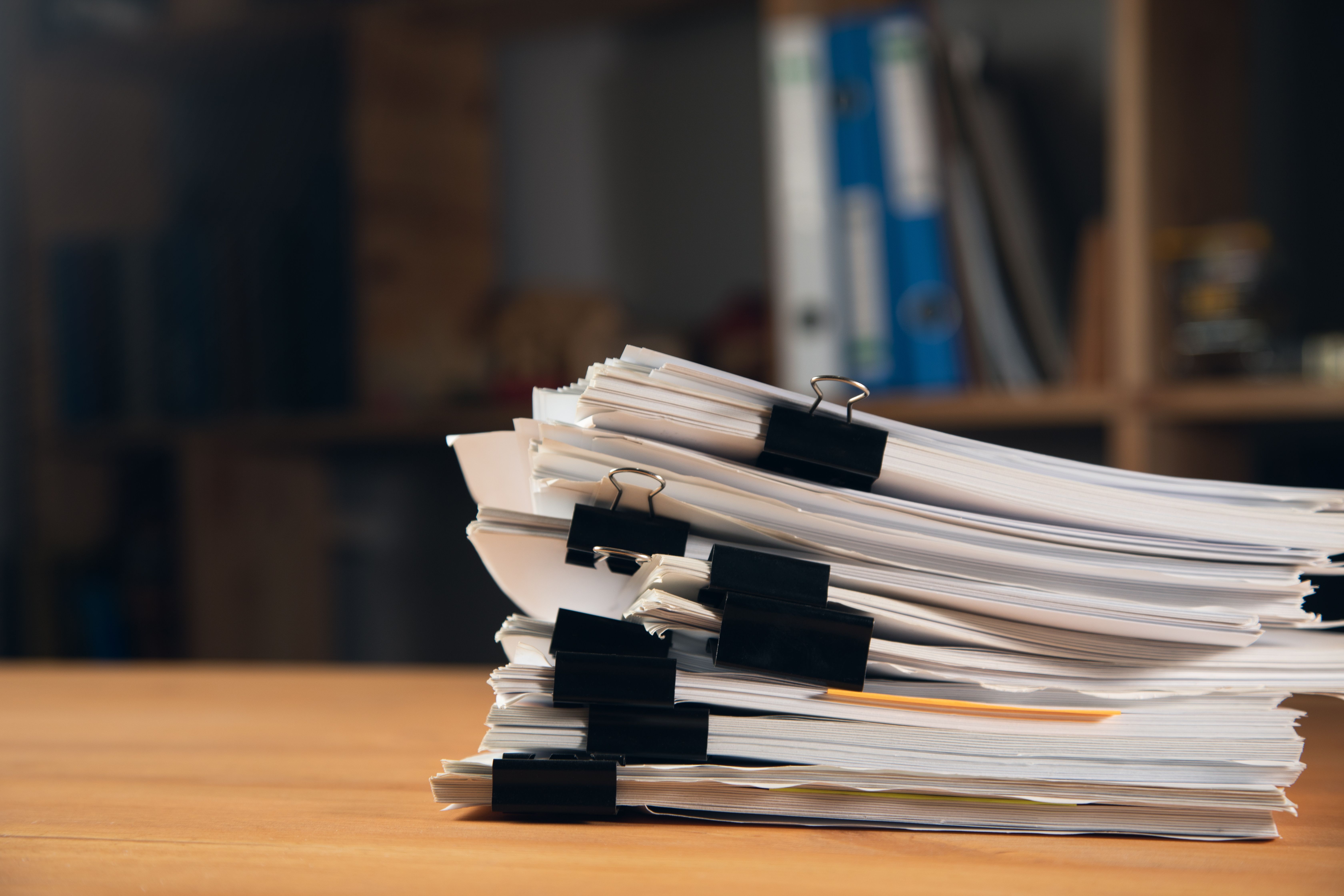 A large stack of printed documents held together with black binder clips sits on a wooden desk, with blurred office shelves in the background.