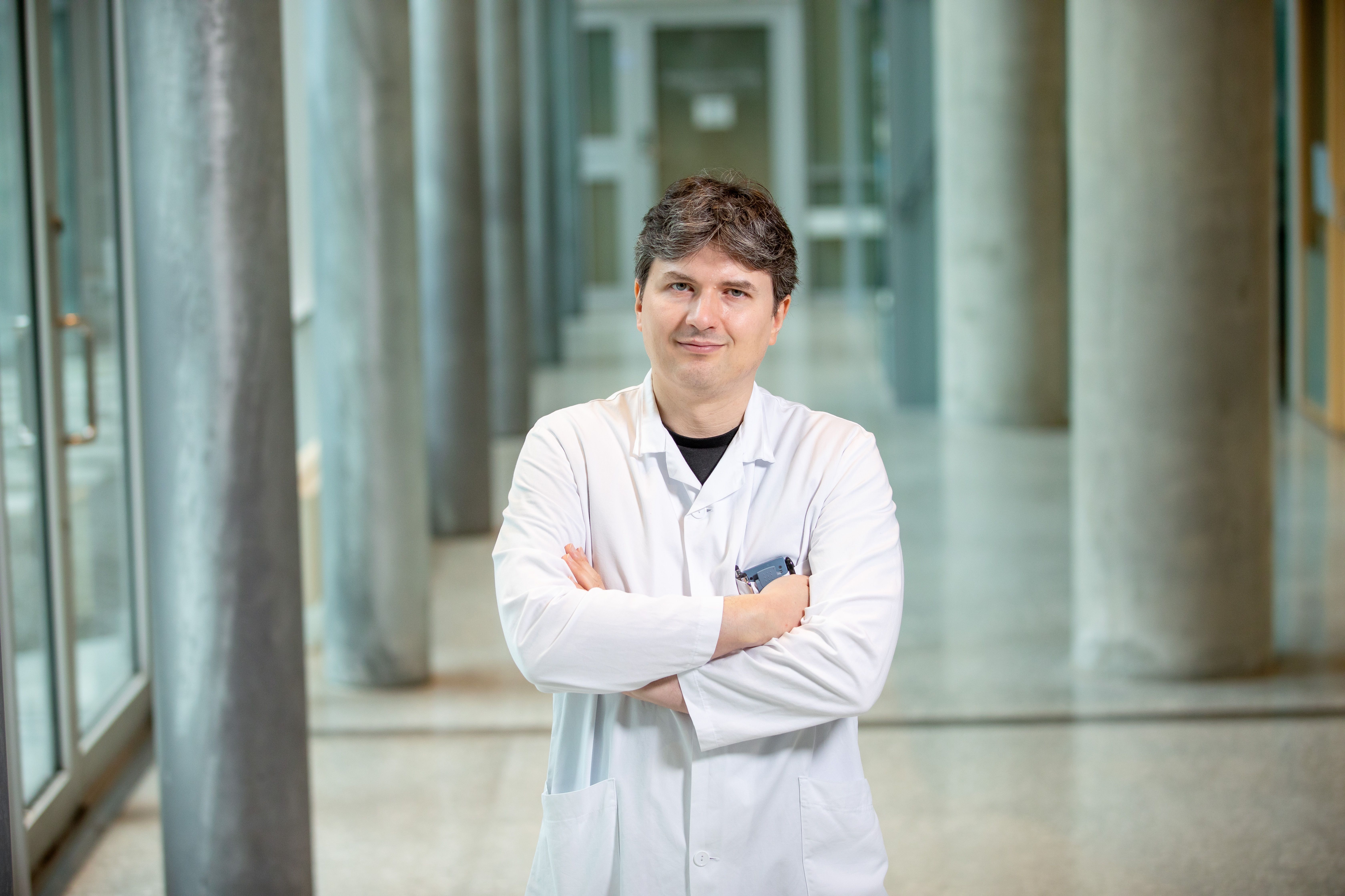 Charalampos Tzoulis, MD, PhD, stands with his arms crossed, wearing a white lab coat in a bright, modern corridor.