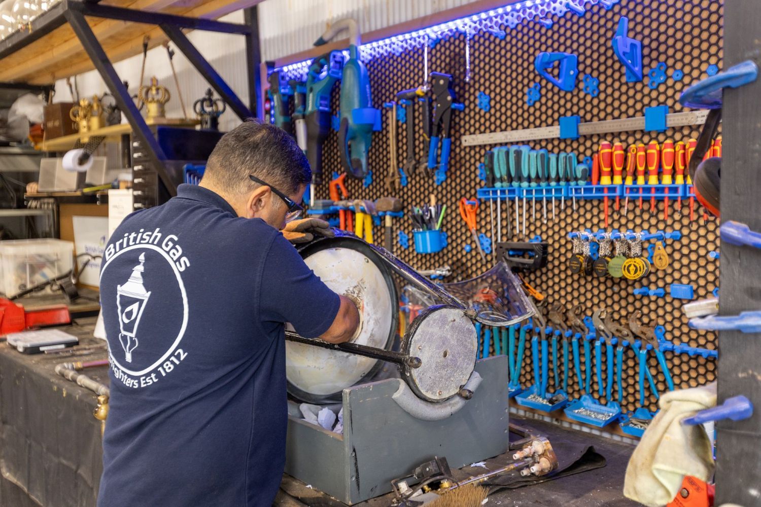 A man in a "British Gas Lamp Lighters" shirt works on an object in a workshop with a tool-filled pegboard wall.