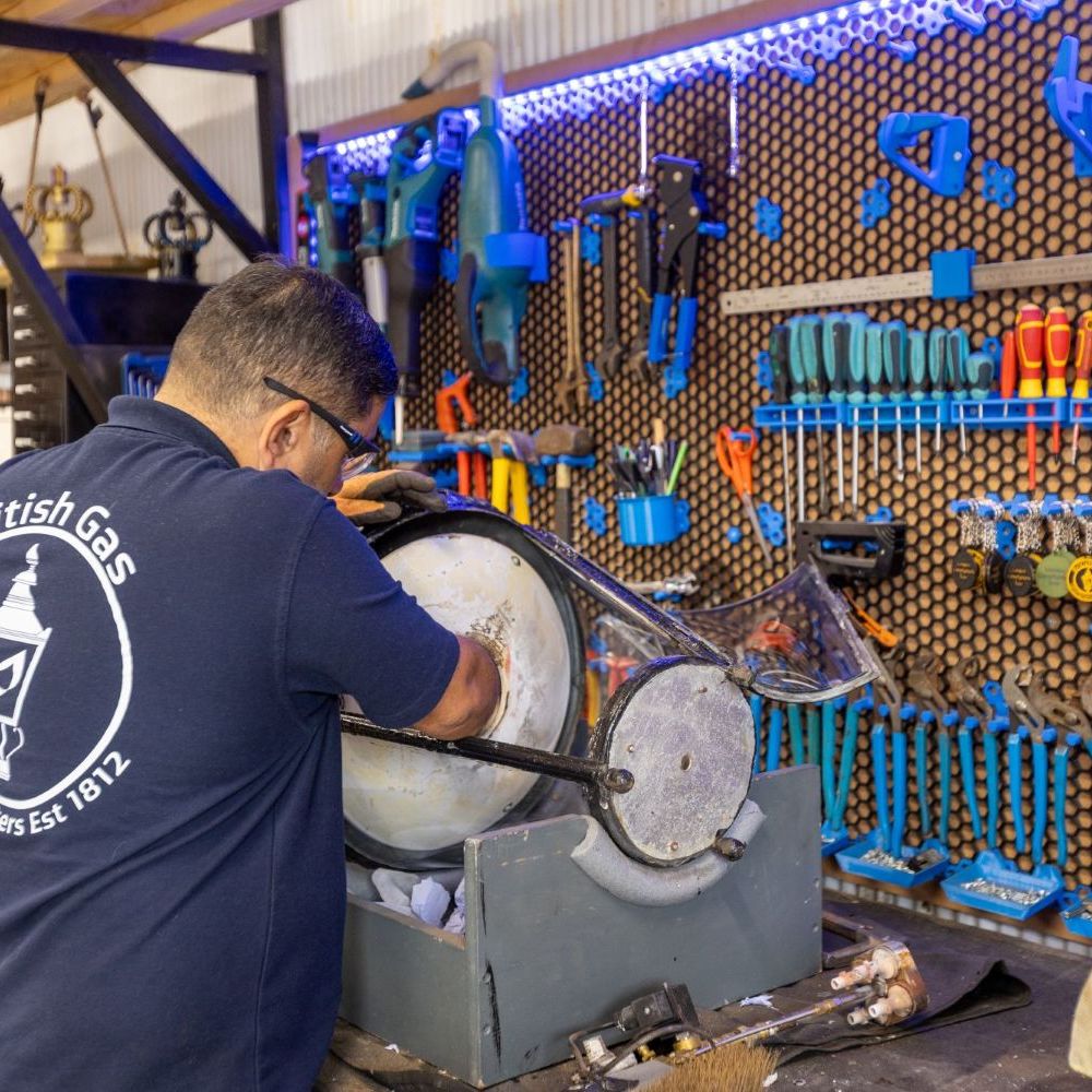 A man in a "British Gas Lamp Lighters" shirt works on an object in a workshop with a tool-filled pegboard wall.