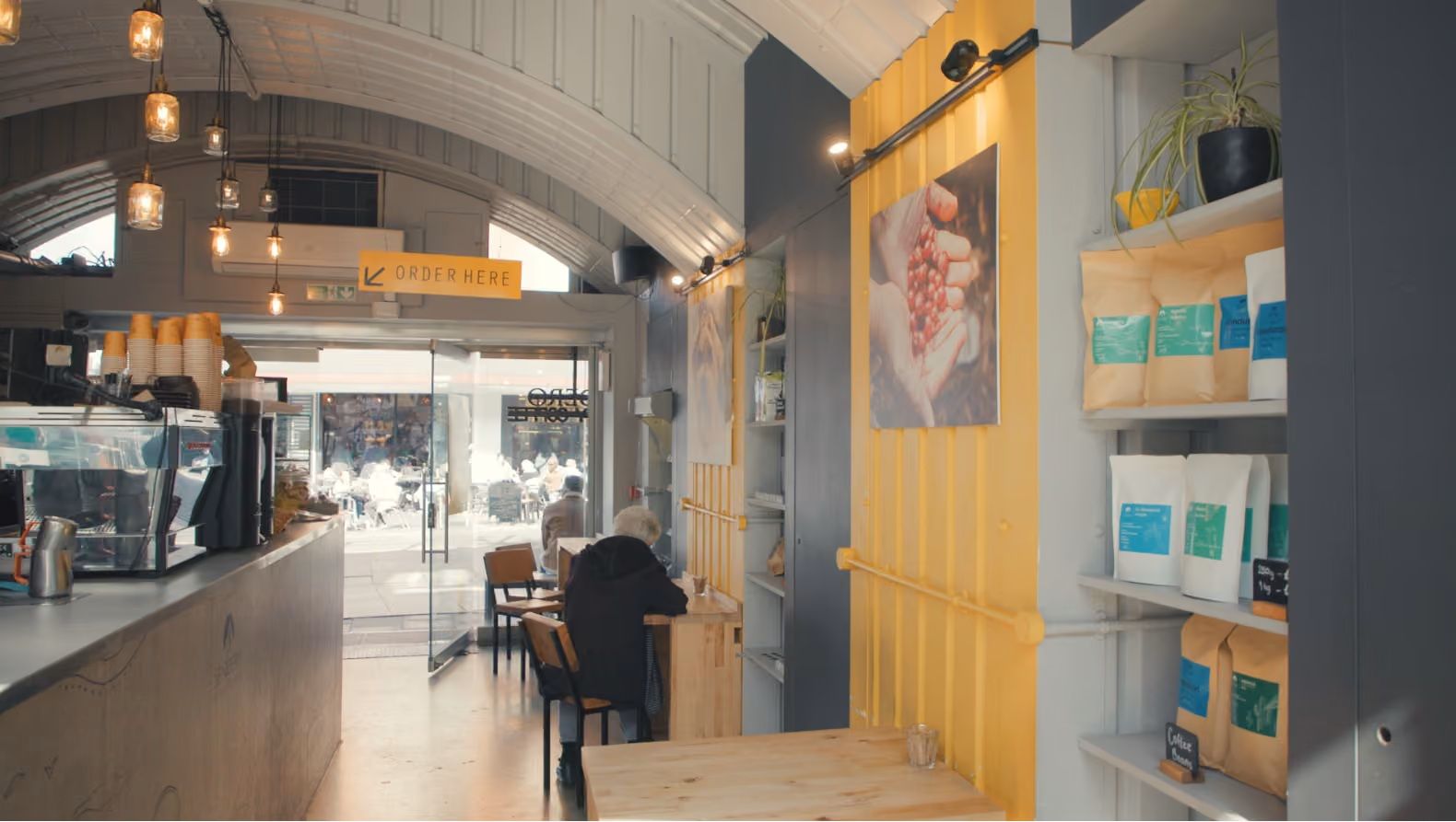 Interior of a modern coffee shop with an arched ceiling, a coffee counter, a customer, and shelves of coffee products.