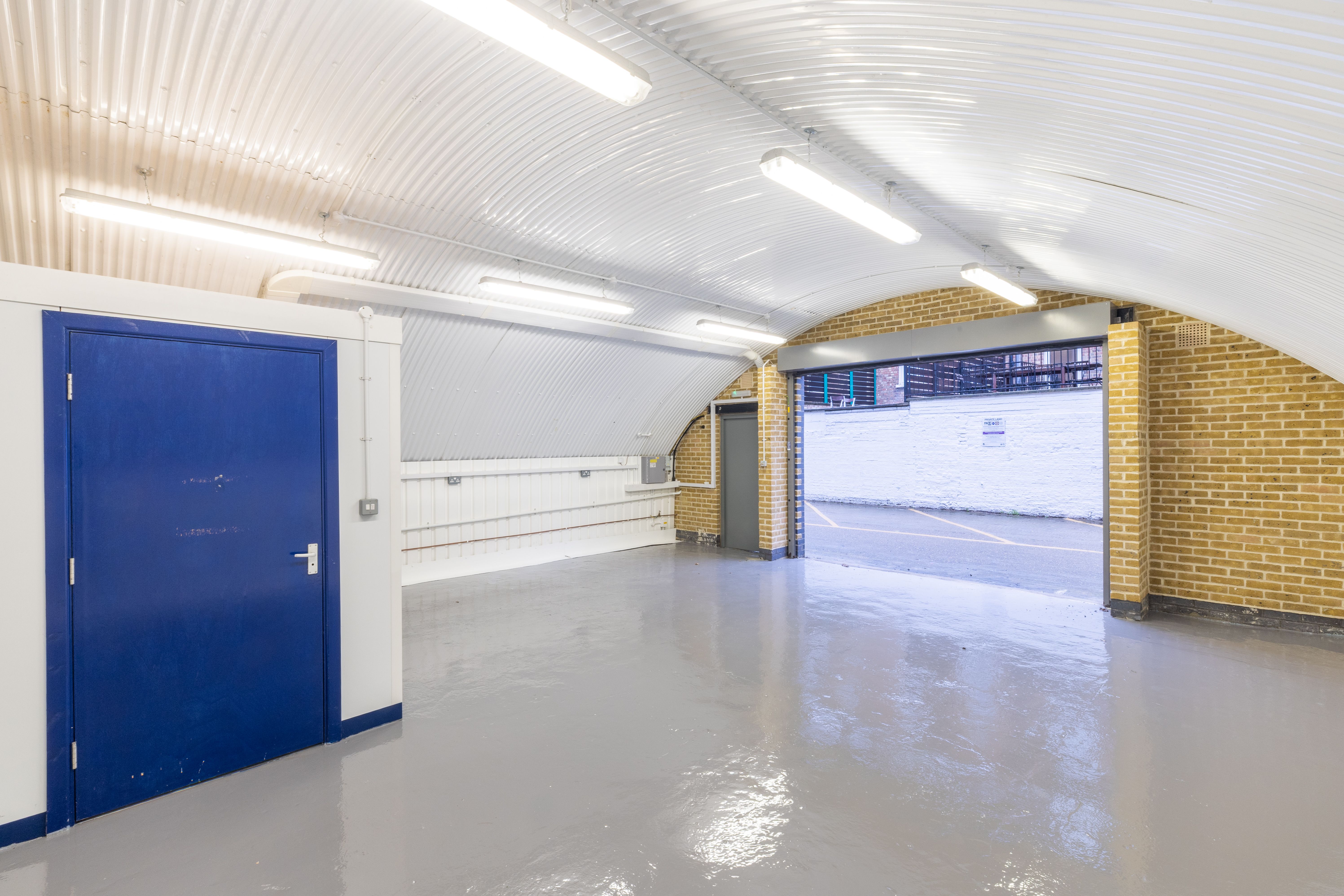 An empty industrial unit with an arched corrugated metal ceiling, gray floor, a blue door on the left, and an open garage door leading outside on the right.