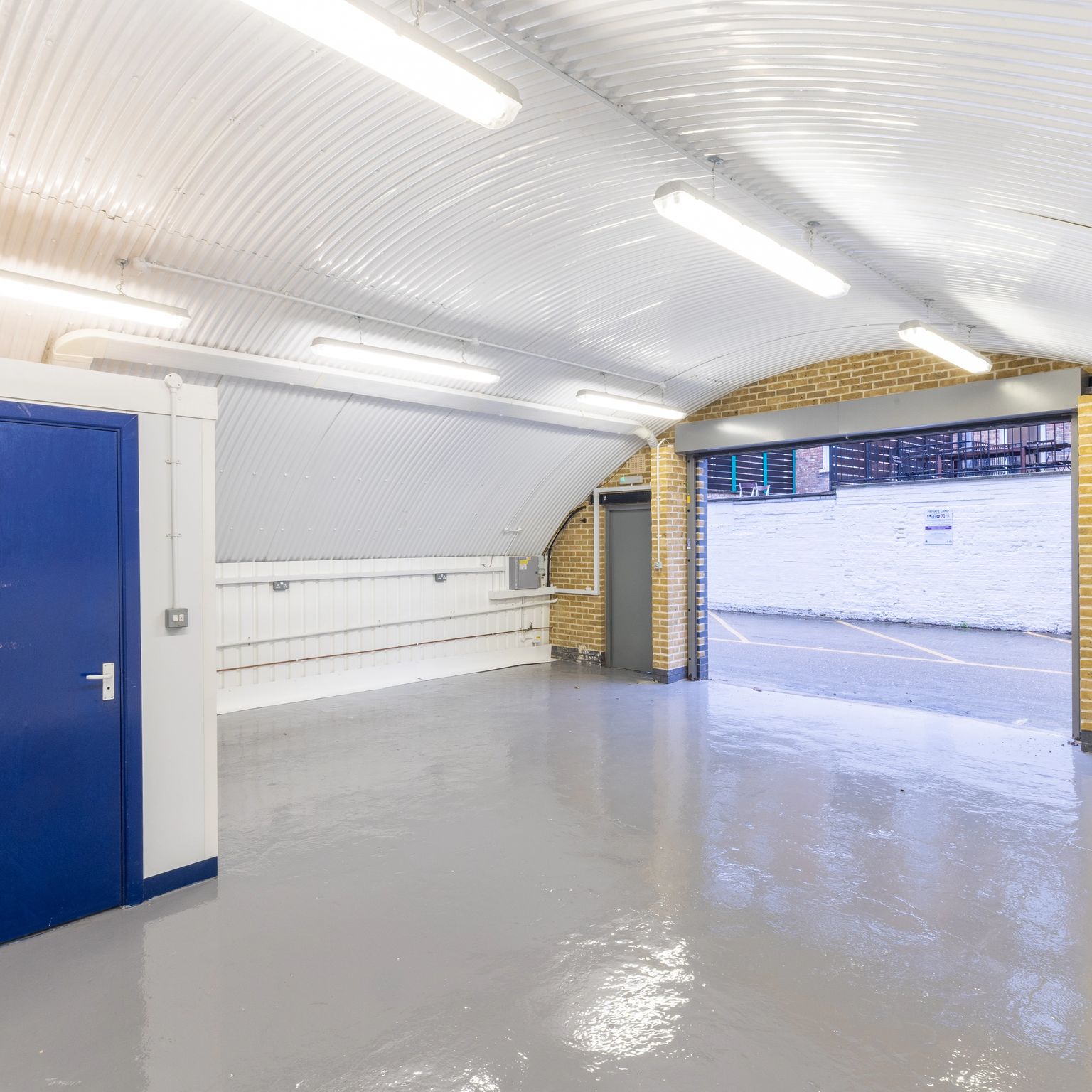 An empty industrial unit with an arched corrugated metal ceiling, gray floor, a blue door on the left, and an open garage door leading outside on the right.