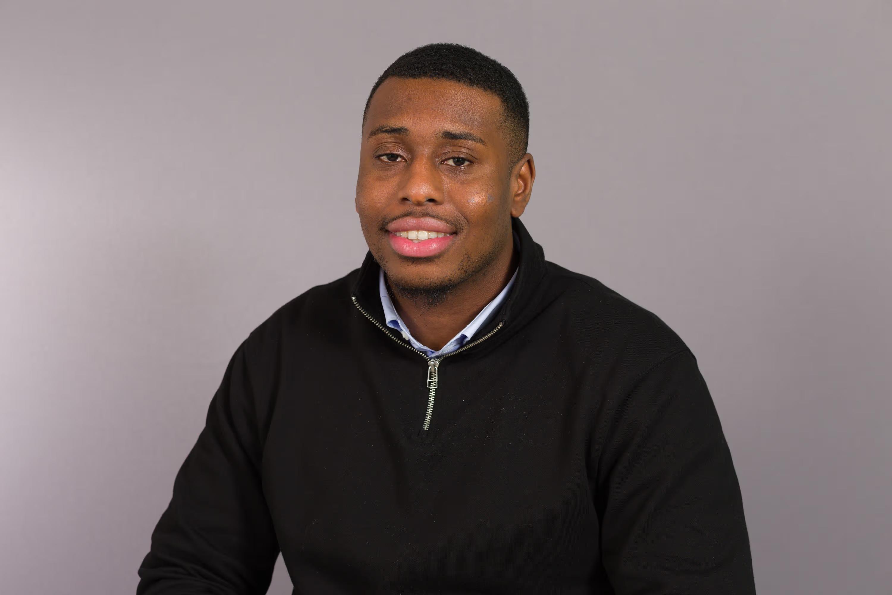 Young Black man smiling in a black quarter-zip pullover against a gray background.