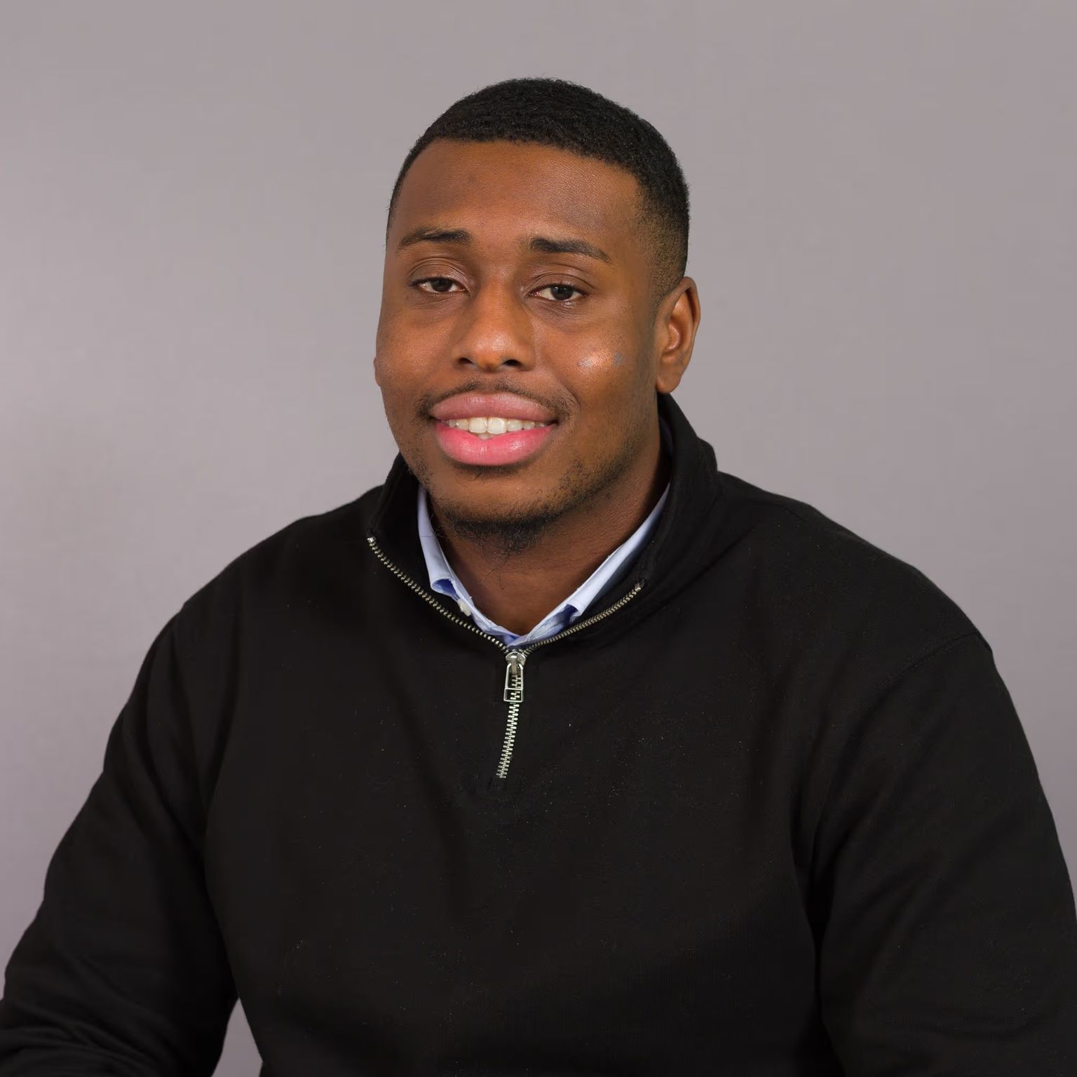Young Black man smiling in a black quarter-zip pullover against a gray background.