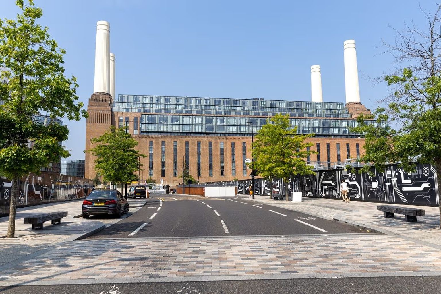 Battersea Power Station, a large brown brick building with four white chimneys and modern glass levels on top, seen from a paved street.