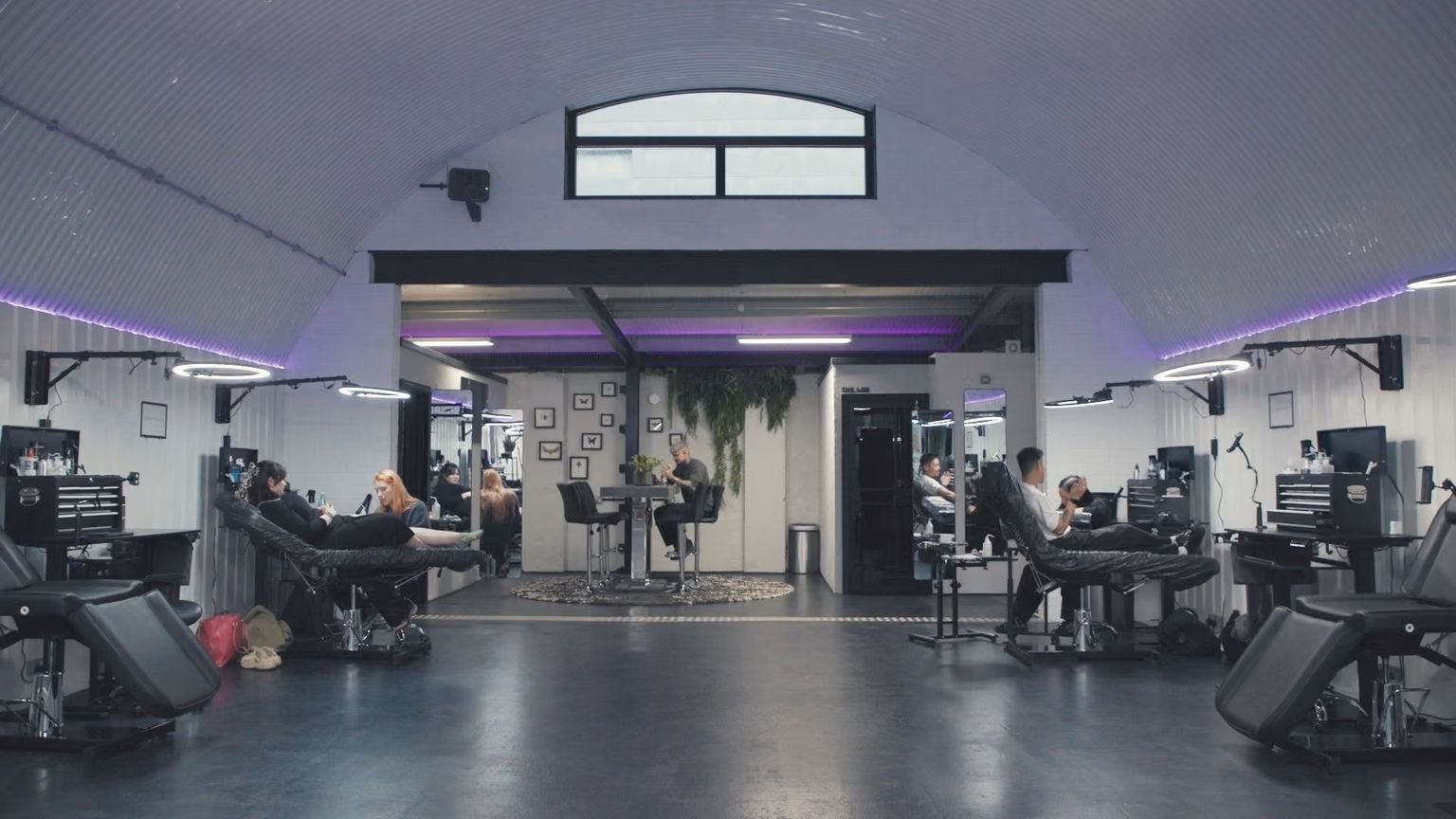 A spacious, modern tattoo studio with multiple stations, clients getting tattoos, and a curved ceiling with purple accent lighting.