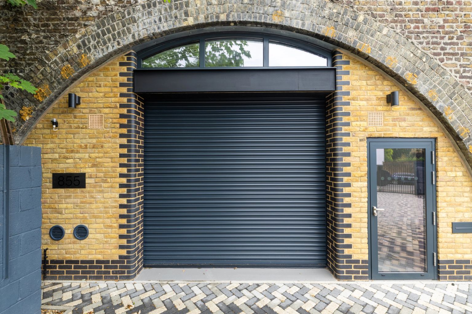 Modern dark grey roller shutter and glass door set into a yellow brick facade within an old brick archway, with the number 855 on the left.