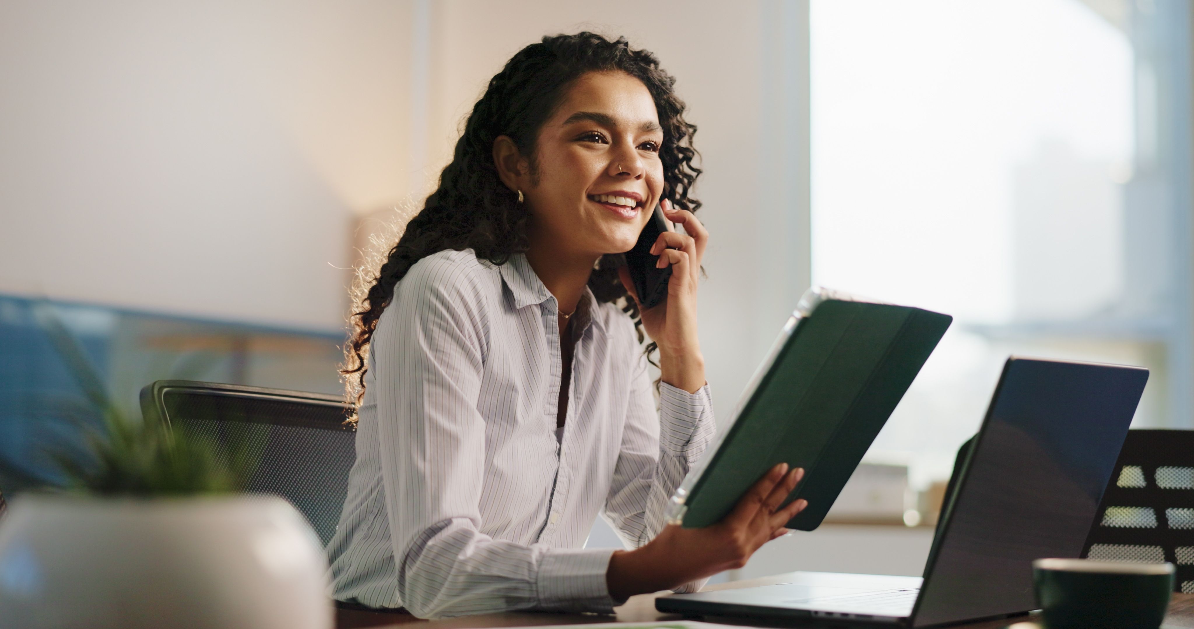 A smiling woman with curly hair talks on a phone, holding a folder at a desk with a laptop.