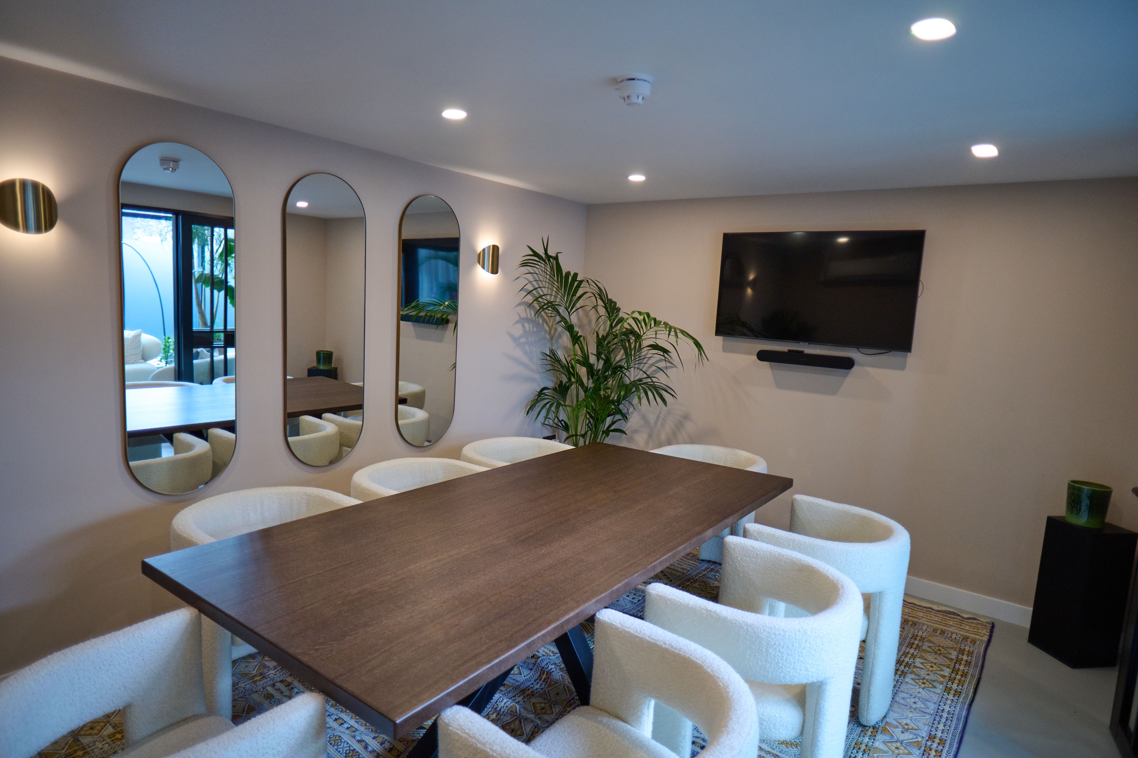 A modern meeting room features a long wooden table, white upholstered chairs, three oval mirrors, a large TV, and a potted plant.