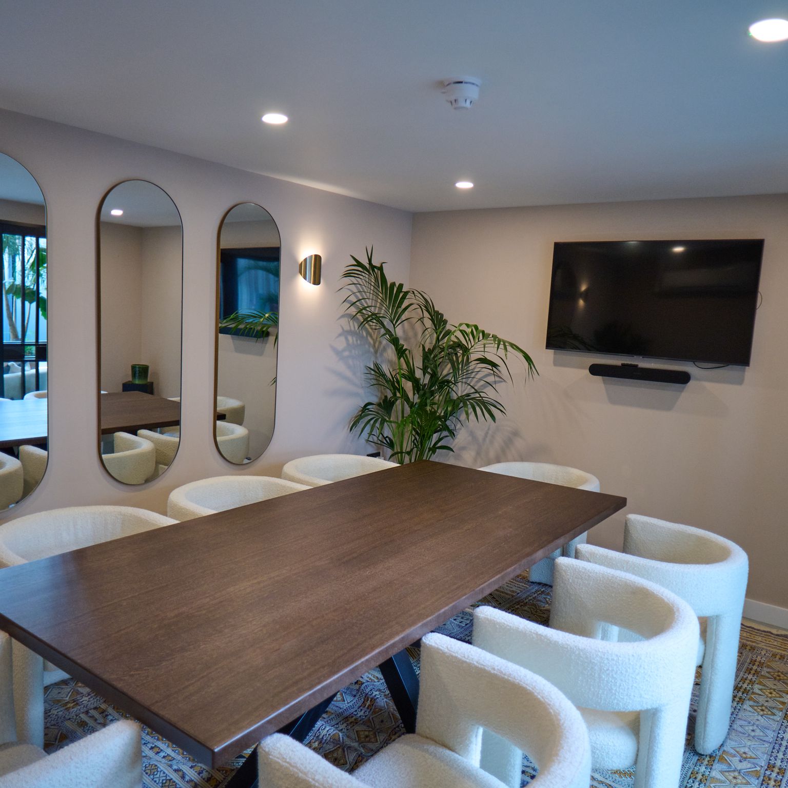 A modern meeting room features a long wooden table, white upholstered chairs, three oval mirrors, a large TV, and a potted plant.