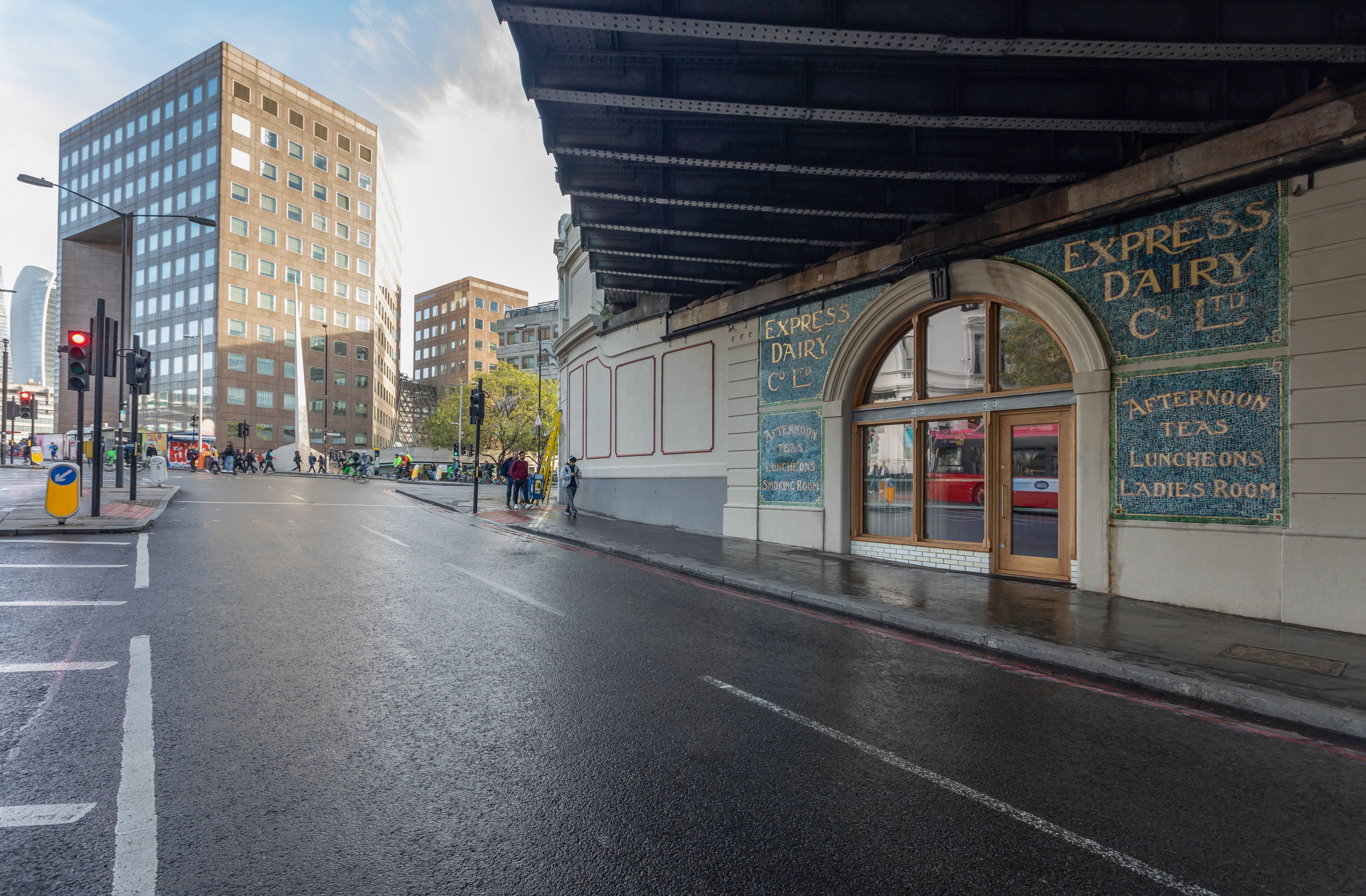 City street with a wet road, a historic "Express Dairy" building under an overhead structure, and modern buildings in the background.