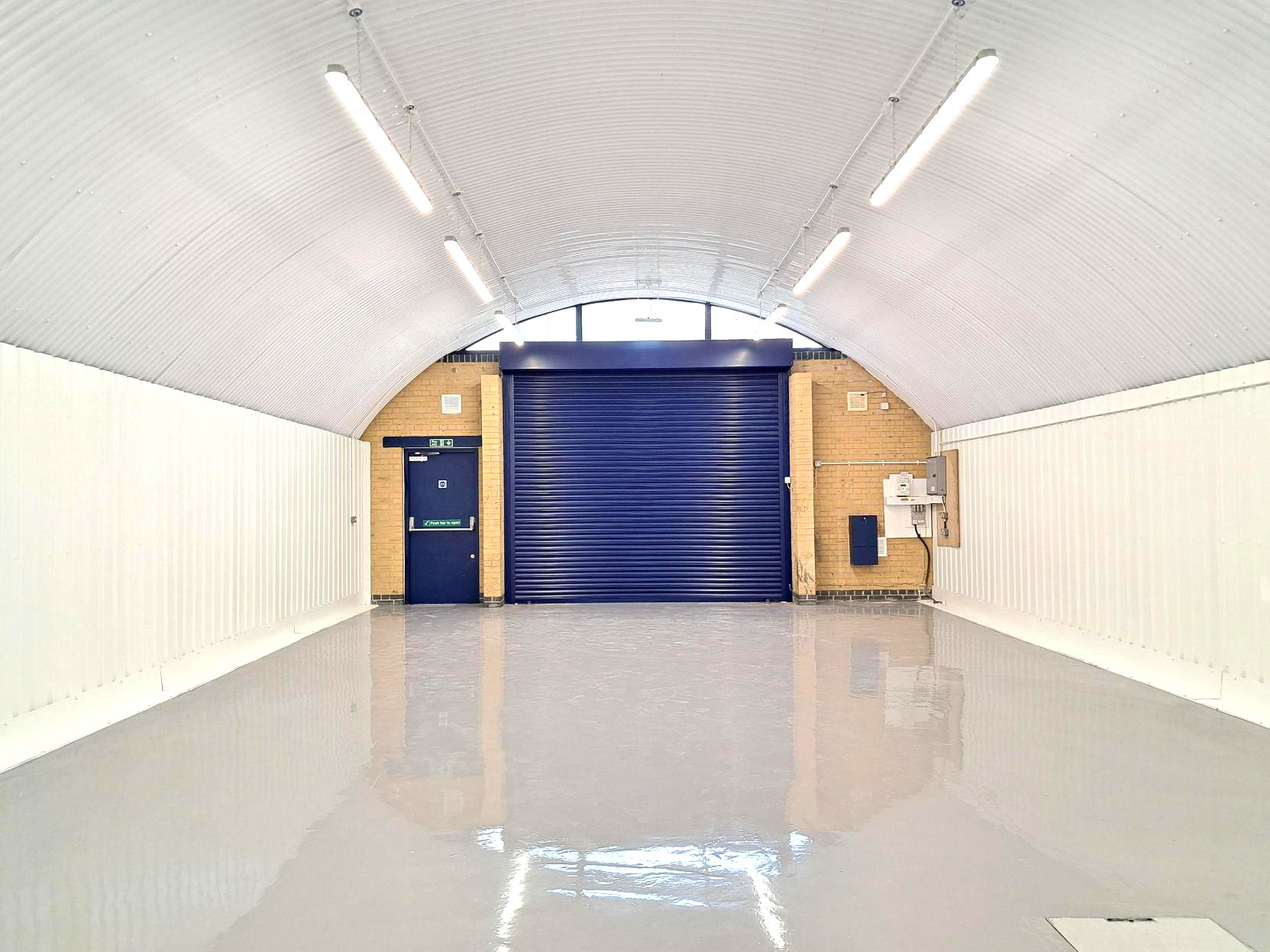 Empty industrial unit with a curved corrugated ceiling, shiny gray floor, and a blue roller door.