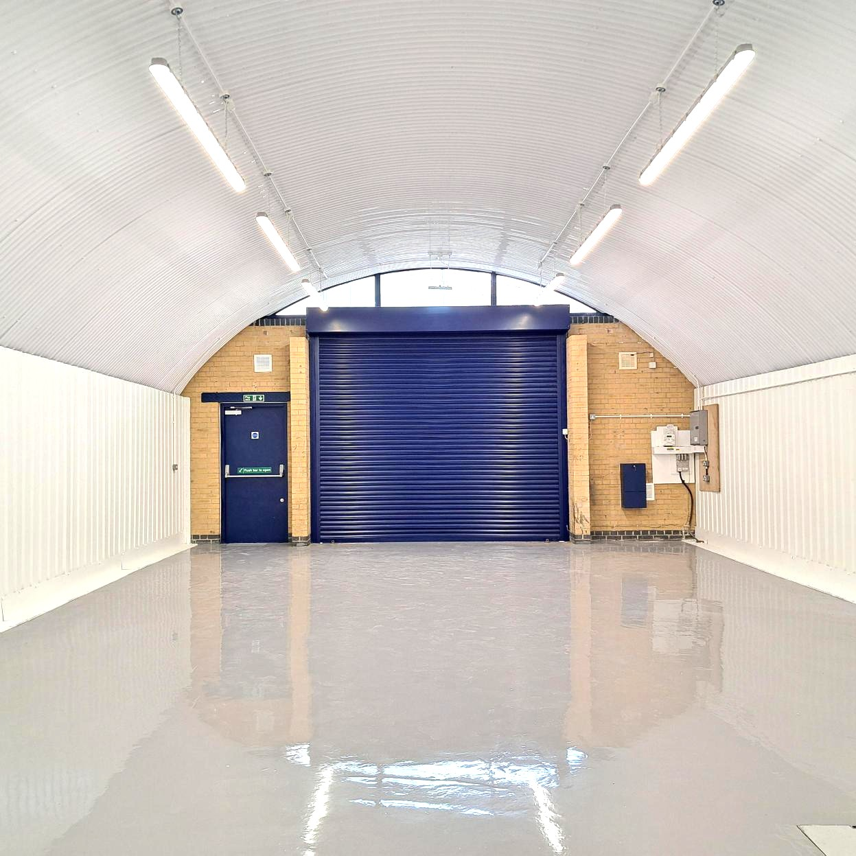 Empty industrial unit with a curved corrugated ceiling, shiny gray floor, and a blue roller door.