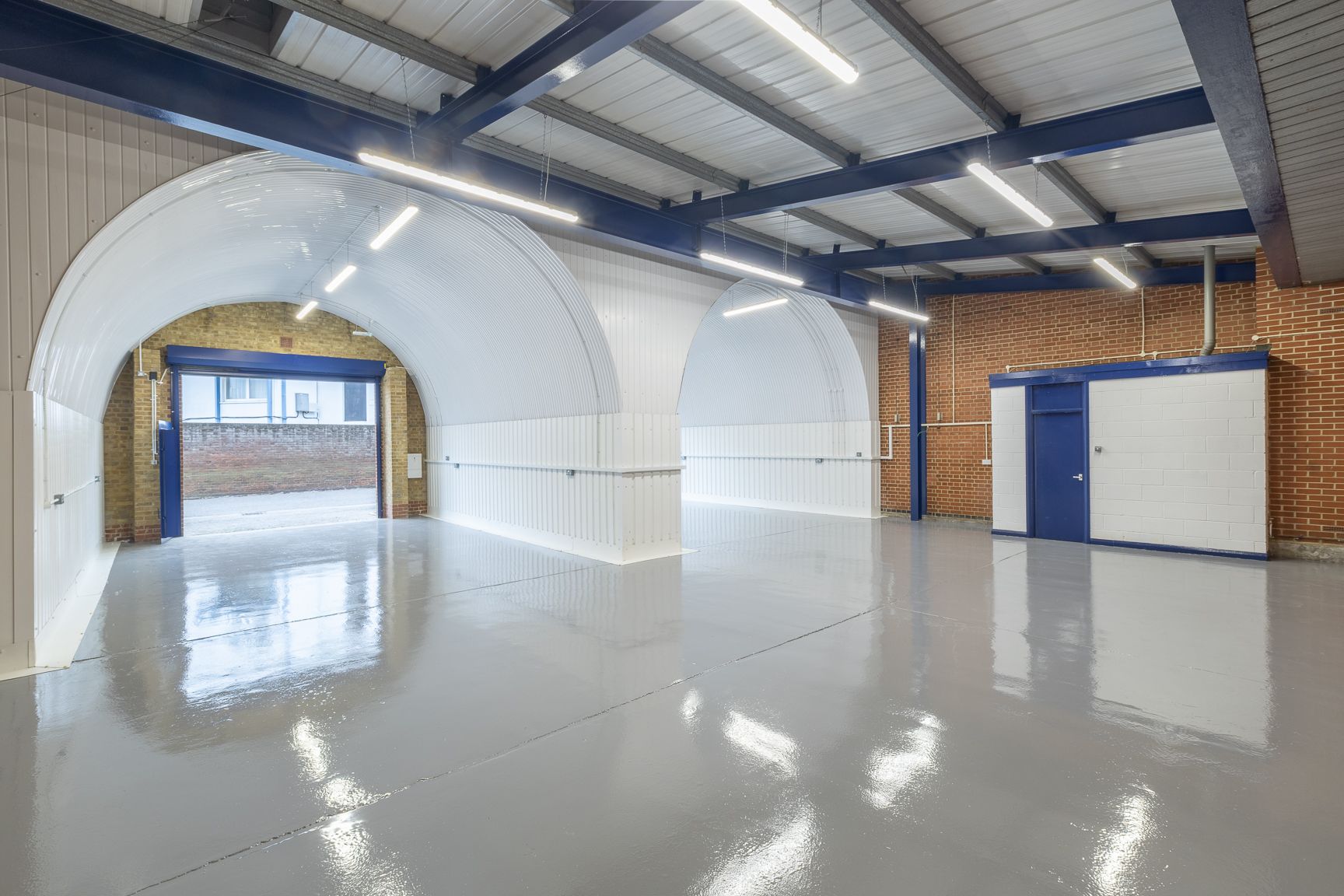 An empty industrial unit with a glossy grey floor, white arched structures, and an open blue roller shutter door.