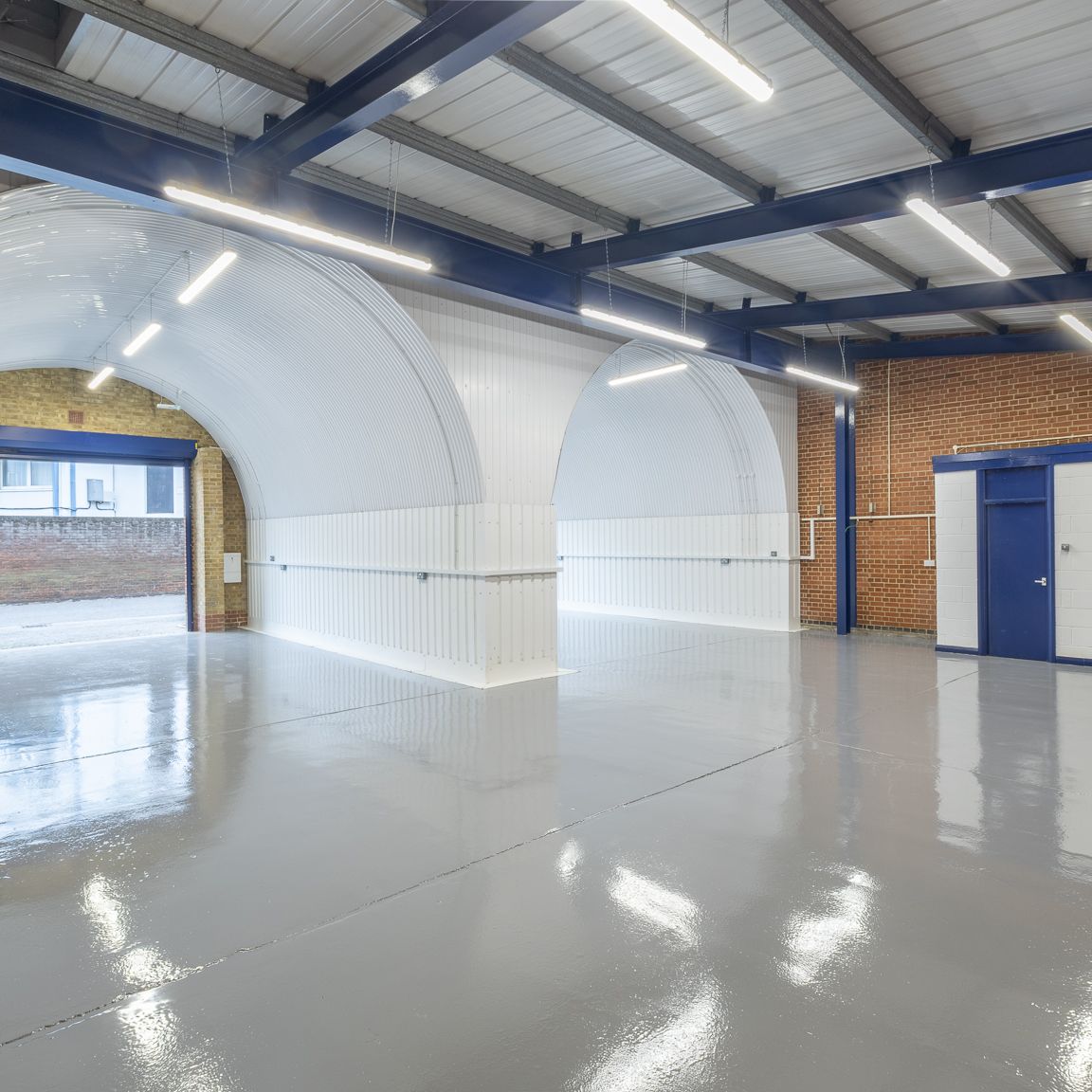 An empty industrial unit with a glossy grey floor, white arched structures, and an open blue roller shutter door.