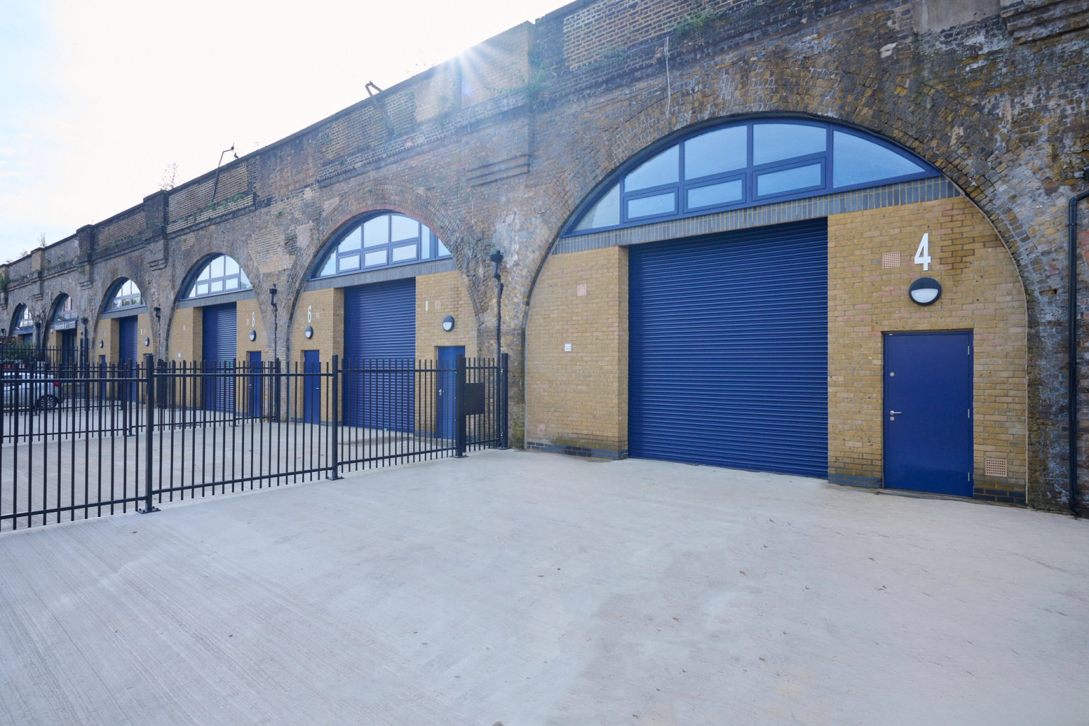 Renovated brick railway arches with blue roll-up doors, arched windows, and a black fence in the foreground.