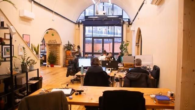 Open-plan office with an arched ceiling, large windows, and several people working at computers.