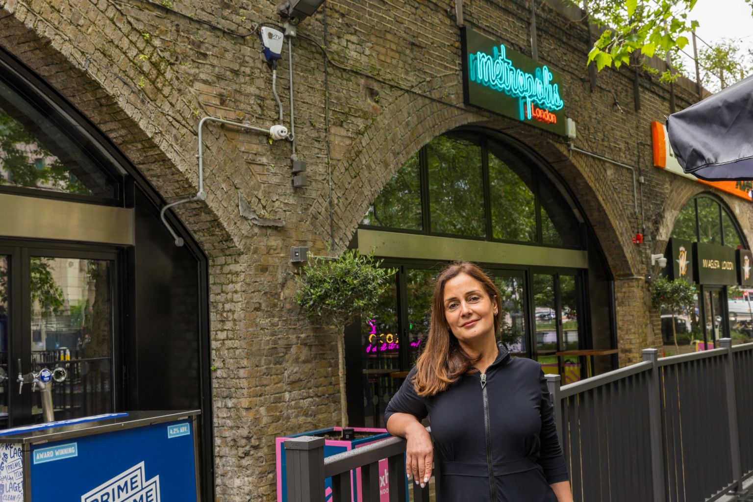 A woman smiles, leaning on a railing outside a brick arch building with a "Metropolis London" sign.