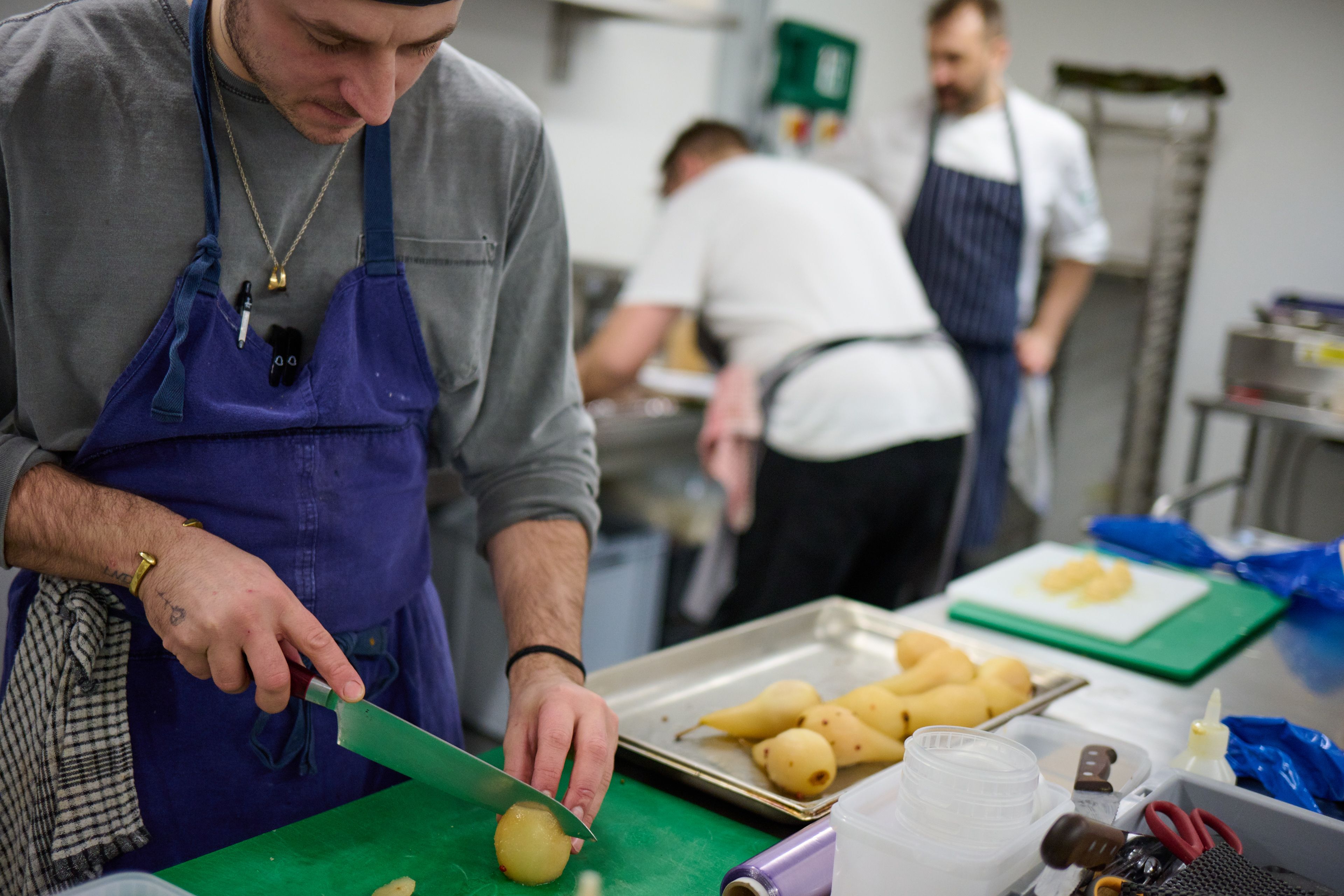 A chef in an apron chops a potato on a green cutting board in a commercial kitchen.