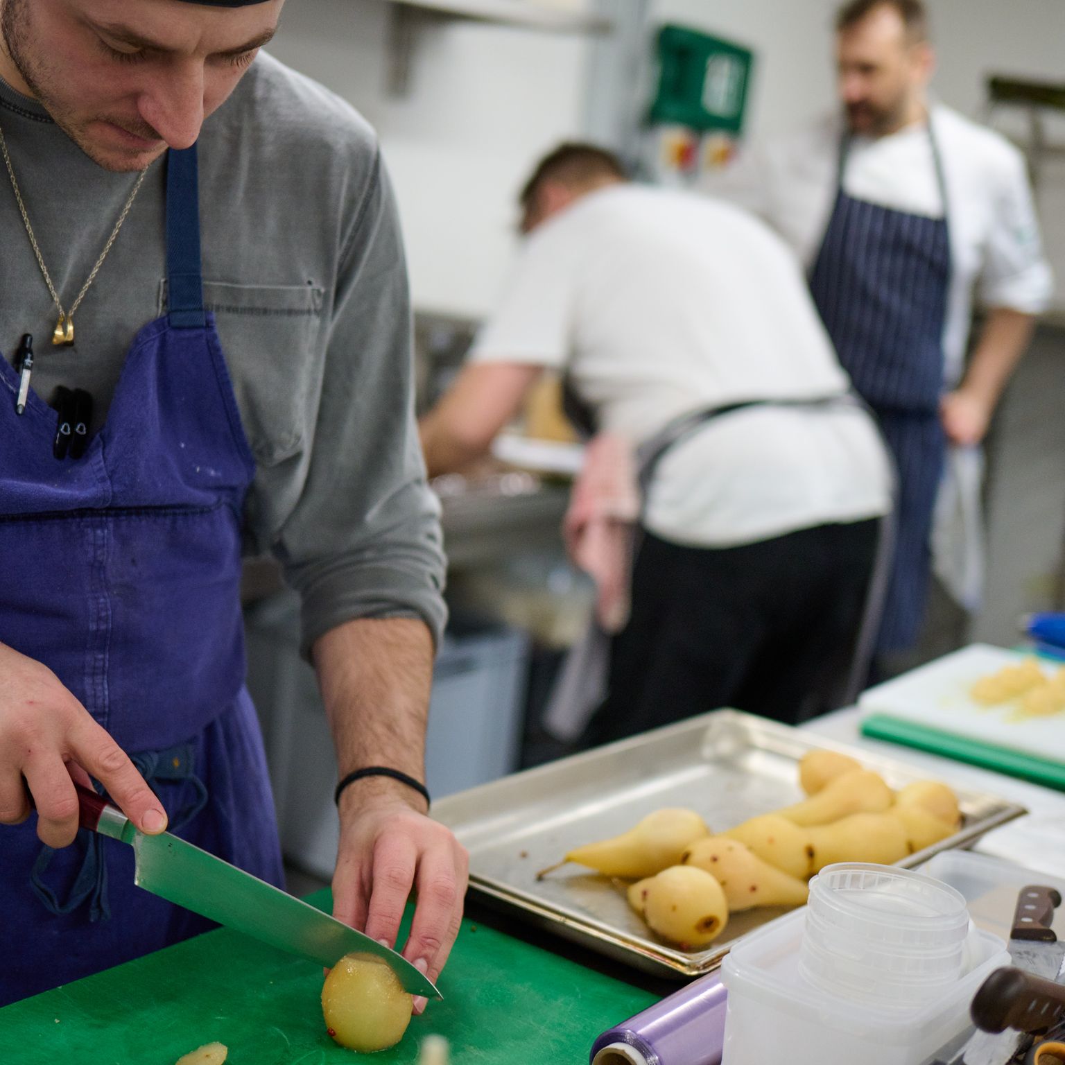 A chef in an apron chops a potato on a green cutting board in a commercial kitchen.