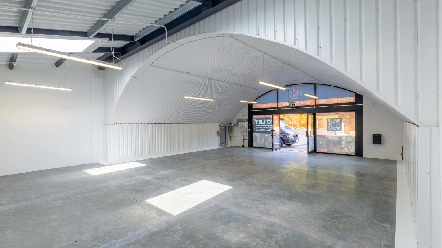 An empty, modern commercial space with a concrete floor, white walls, and an arched entrance with glass doors.