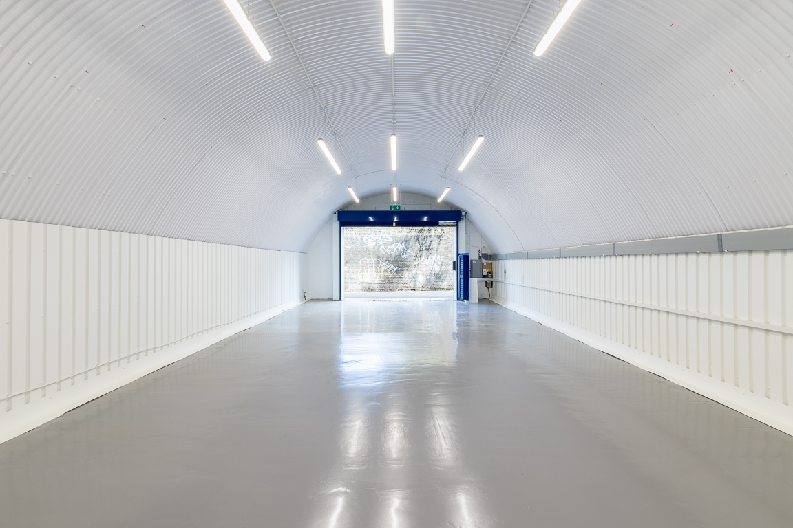Empty white corrugated quonset hut interior with a grey floor and open blue door leading to an outdoor view.