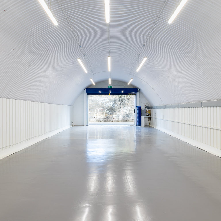 Empty white corrugated quonset hut interior with a grey floor and open blue door leading to an outdoor view.