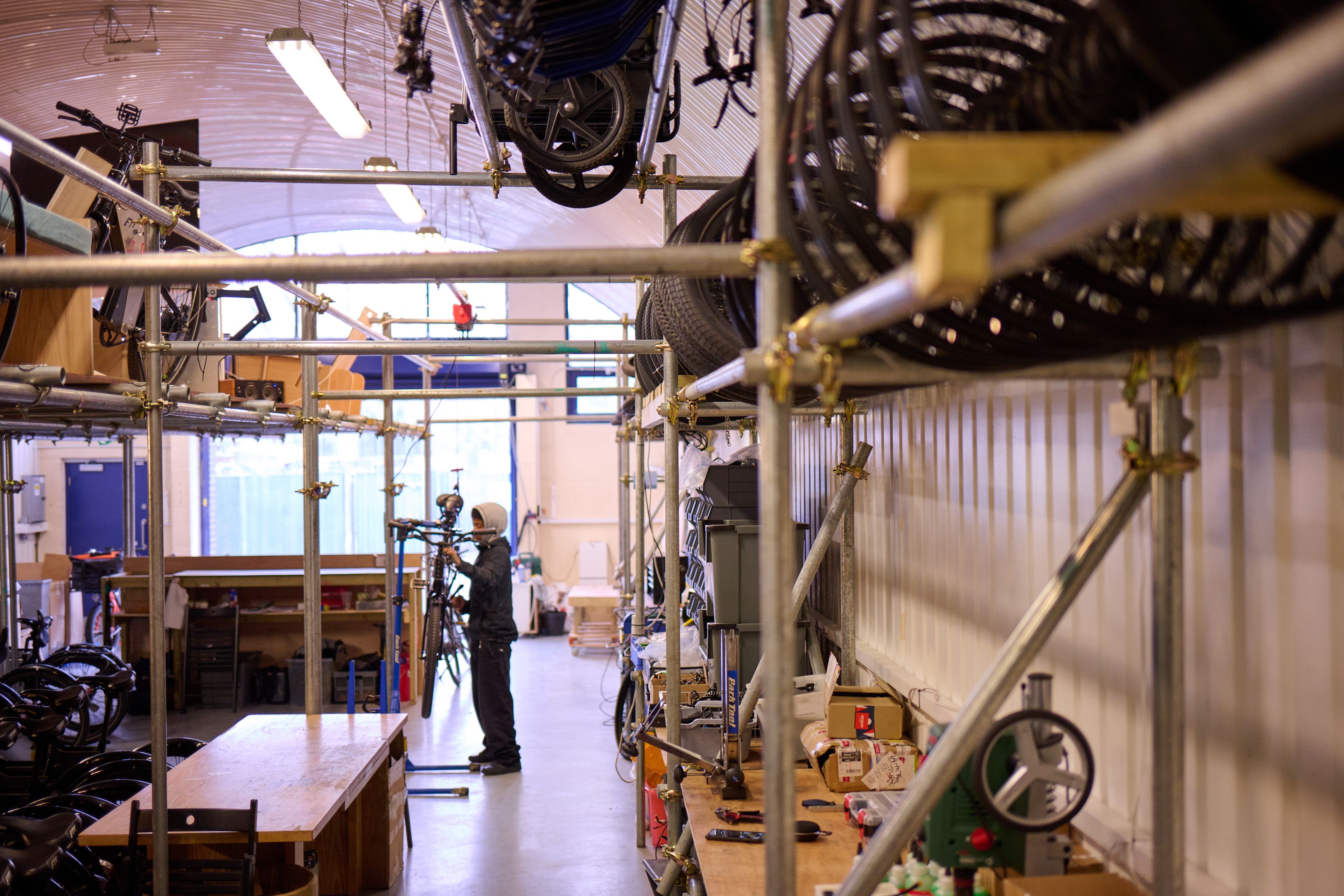 A person repairs a bicycle in a workshop with bikes stored on tiered metal racks, under an arched ceiling.