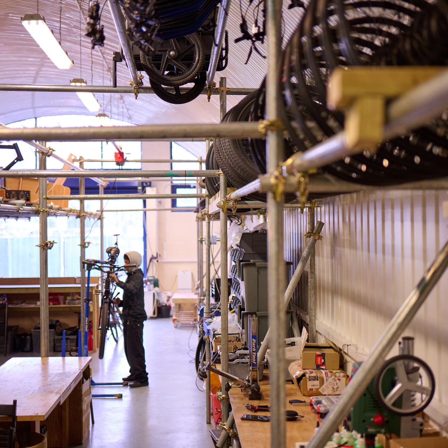 A person repairs a bicycle in a workshop with bikes stored on tiered metal racks, under an arched ceiling.