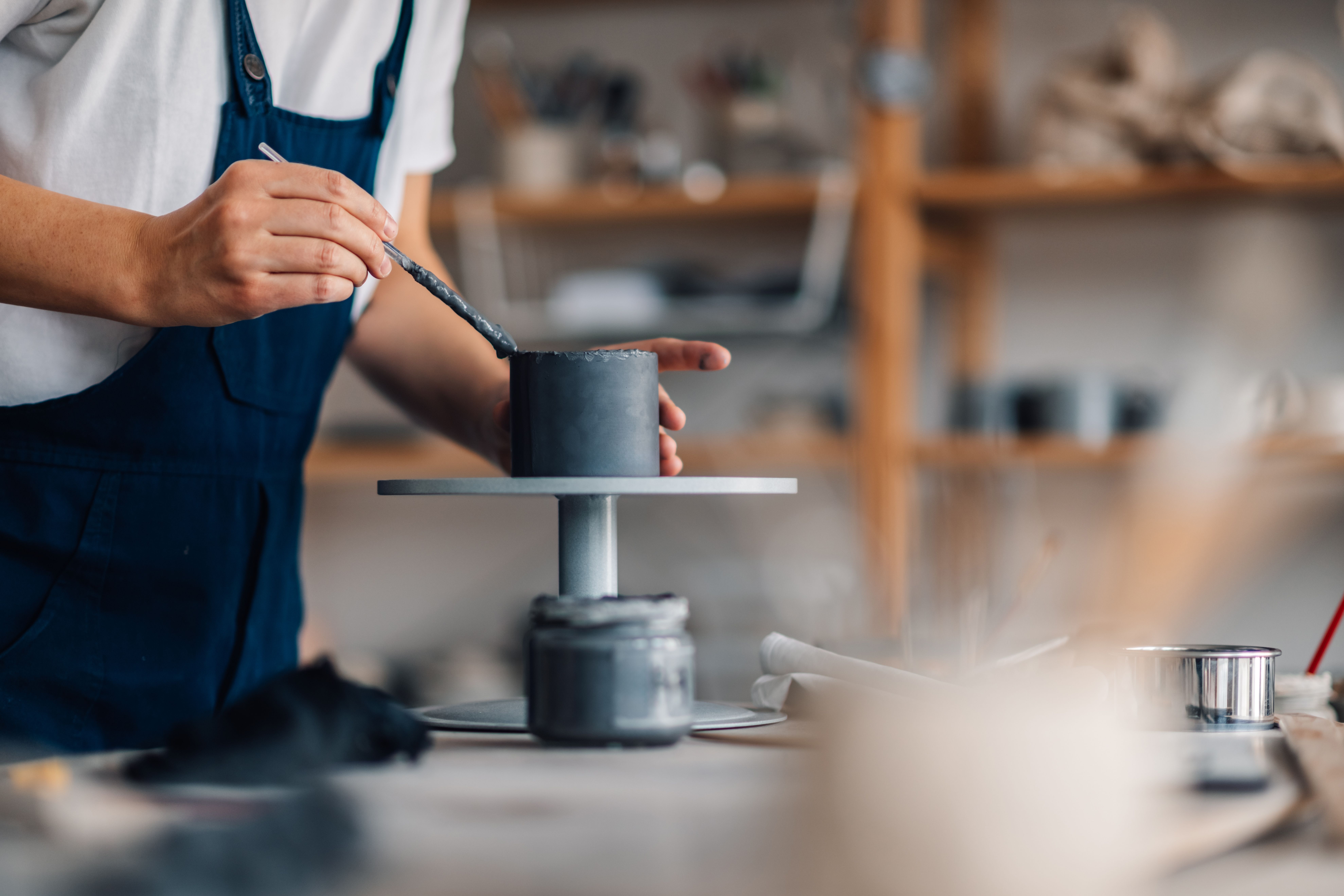 A person in blue overalls glazes a gray ceramic pot with a brush.