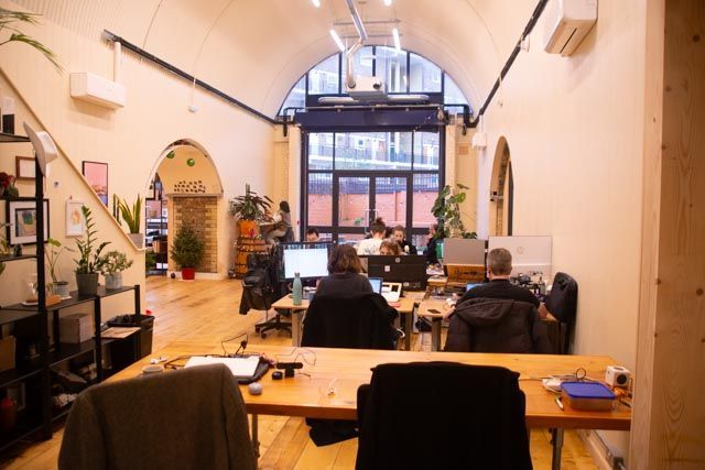 Open-plan office with arched architecture, wooden floors, plants, and several people working at computers.