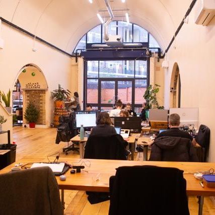 Open-plan office with arched architecture, wooden floors, plants, and several people working at computers.