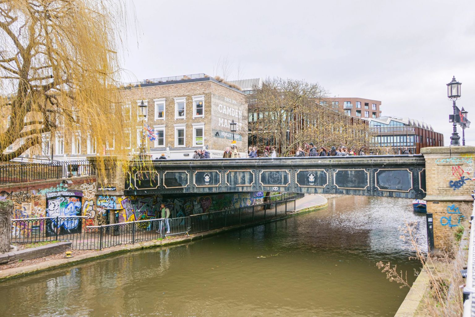 A canal flows under a bridge crowded with people in Camden Market, flanked by graffiti-covered banks and a weeping willow tree.