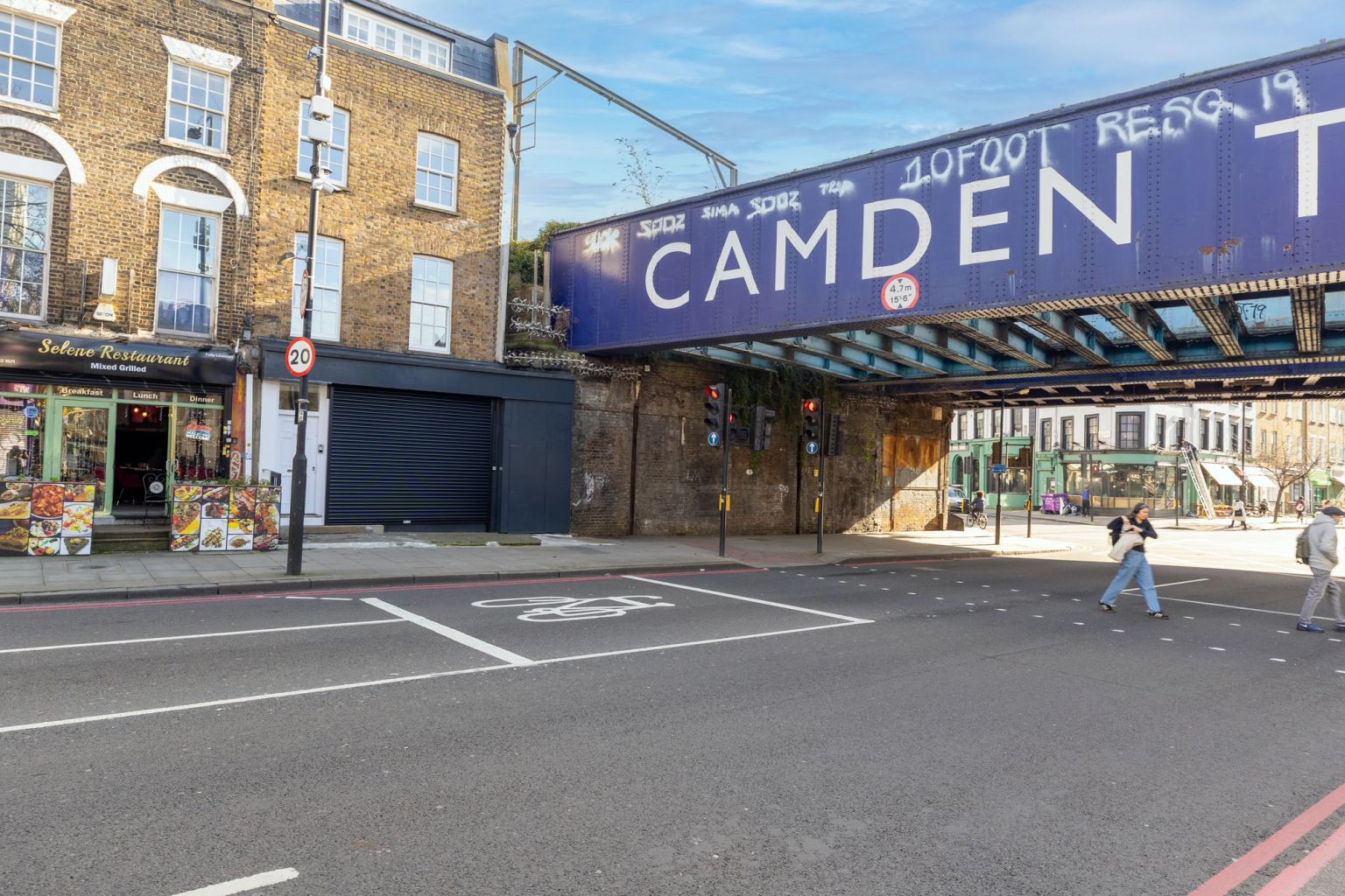 Camden street scene with a blue railway bridge reading "CAMDEN" over a road with pedestrians and brick buildings.