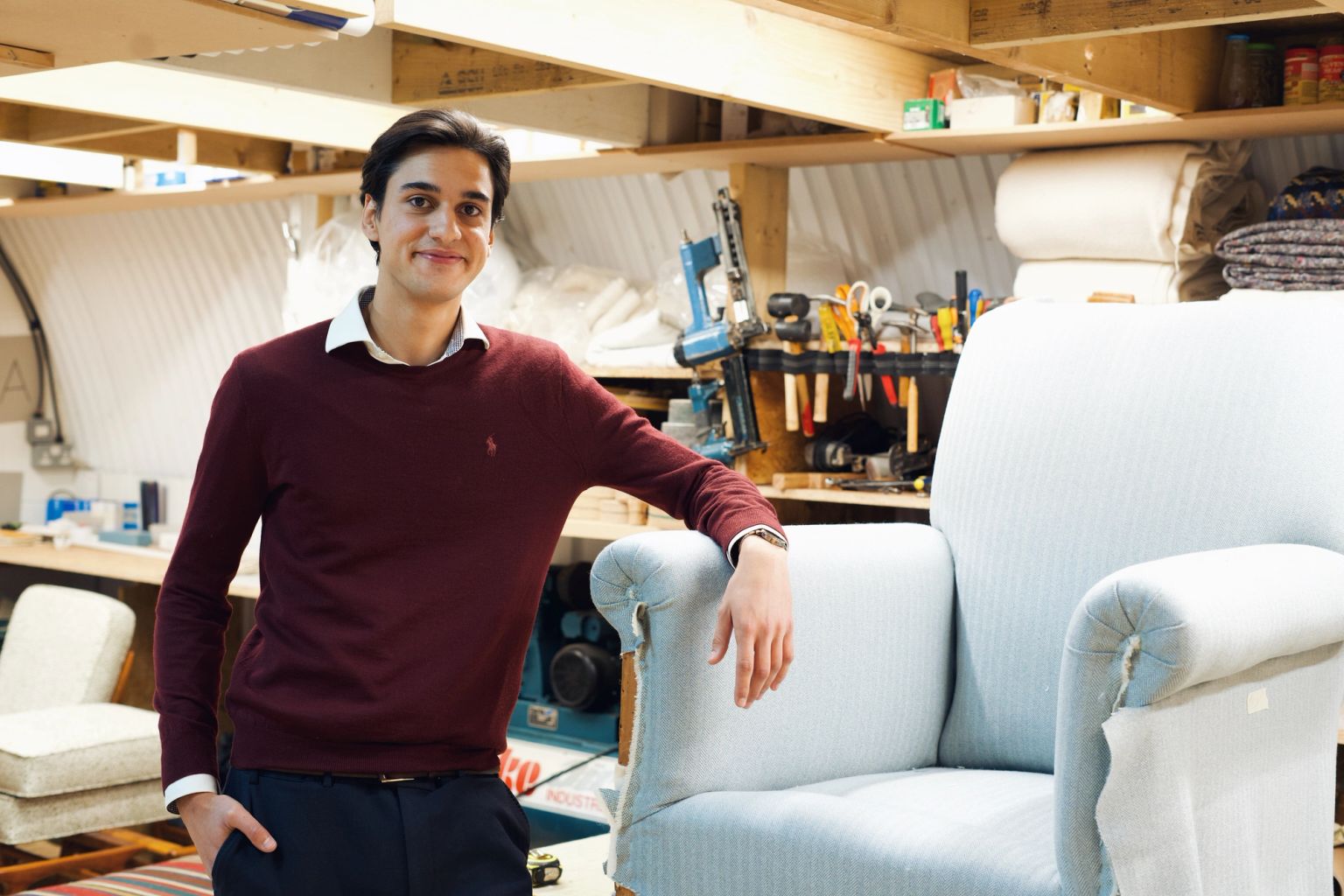 A smiling young man in a workshop, leaning on a partially upholstered light blue armchair.