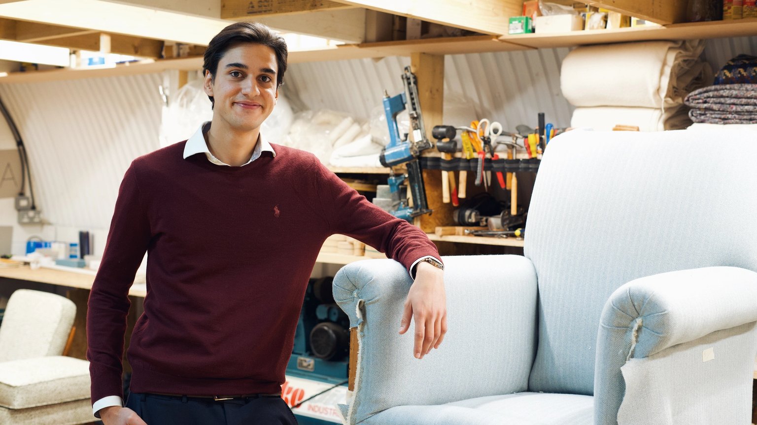 A smiling young man in a workshop, leaning on a partially upholstered light blue armchair.