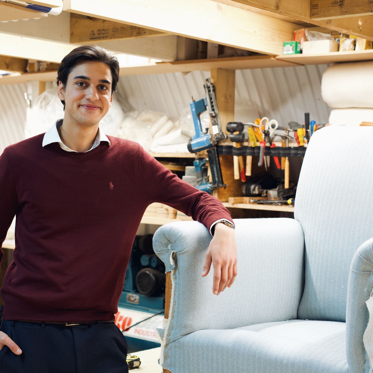 Young man smiling in a workshop, leaning on an armchair being reupholstered.