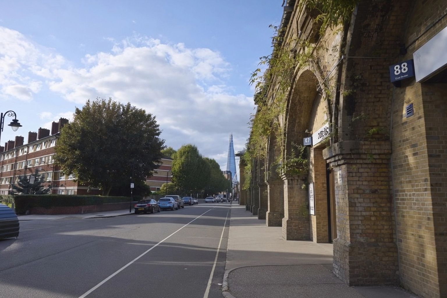 A street scene with brick railway arches on the right, residential buildings on the left, and The Shard skyscraper in the distance under a partly cloudy sky.