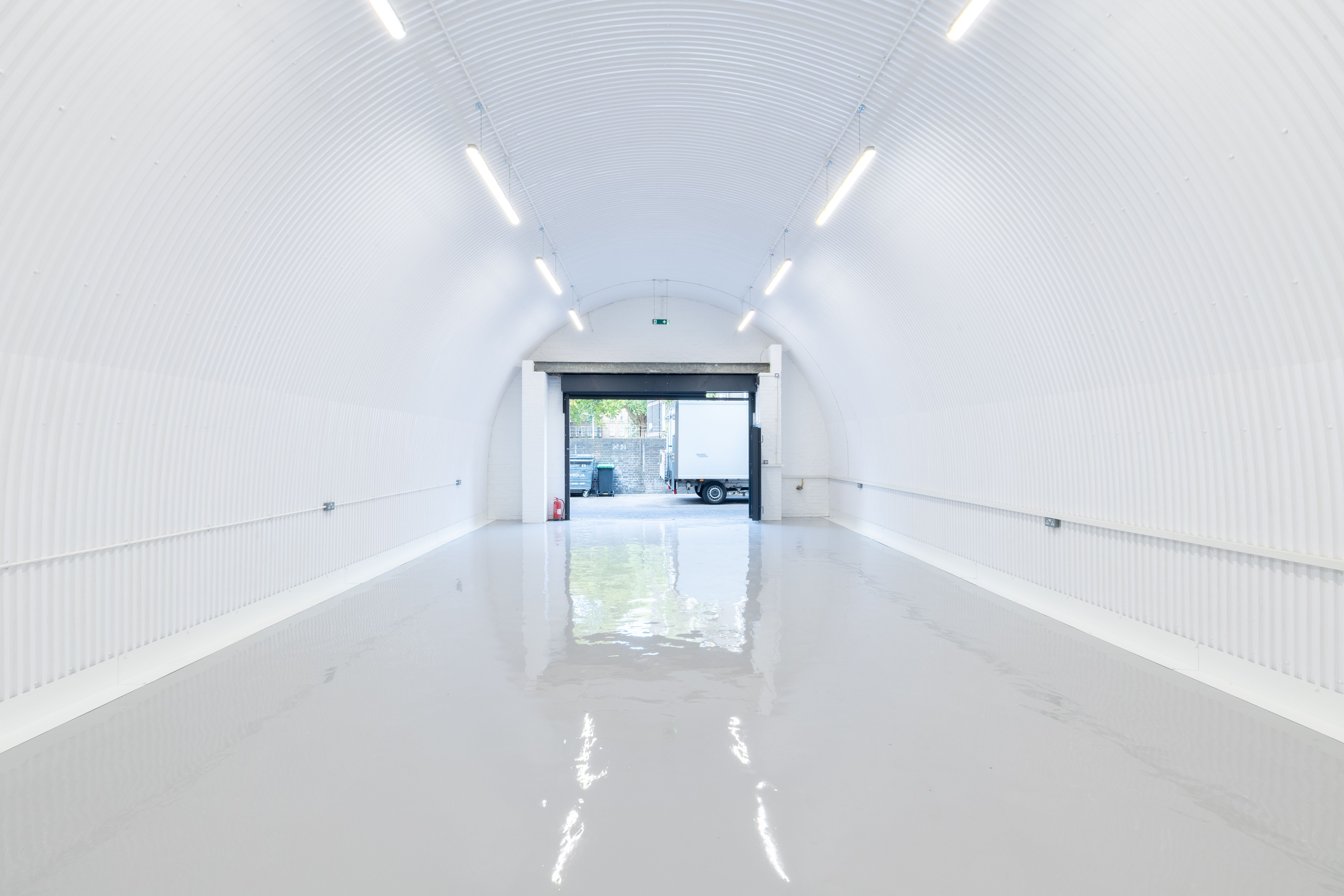 White corrugated arched tunnel with a reflective grey floor, leading to an open bay door with a white truck visible outside.