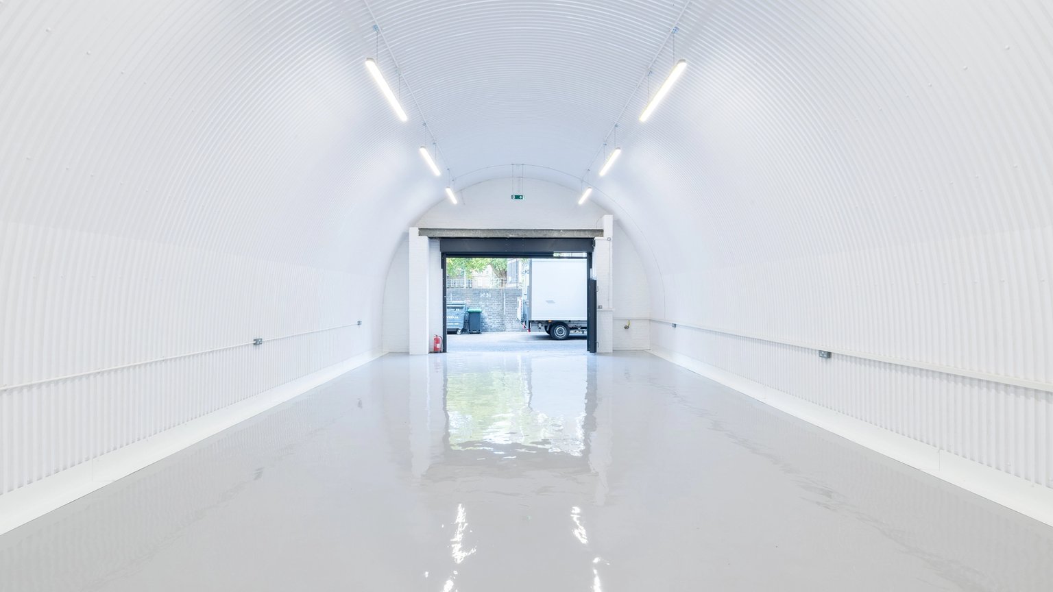 White corrugated arched tunnel with a reflective grey floor, leading to an open bay door with a white truck visible outside.