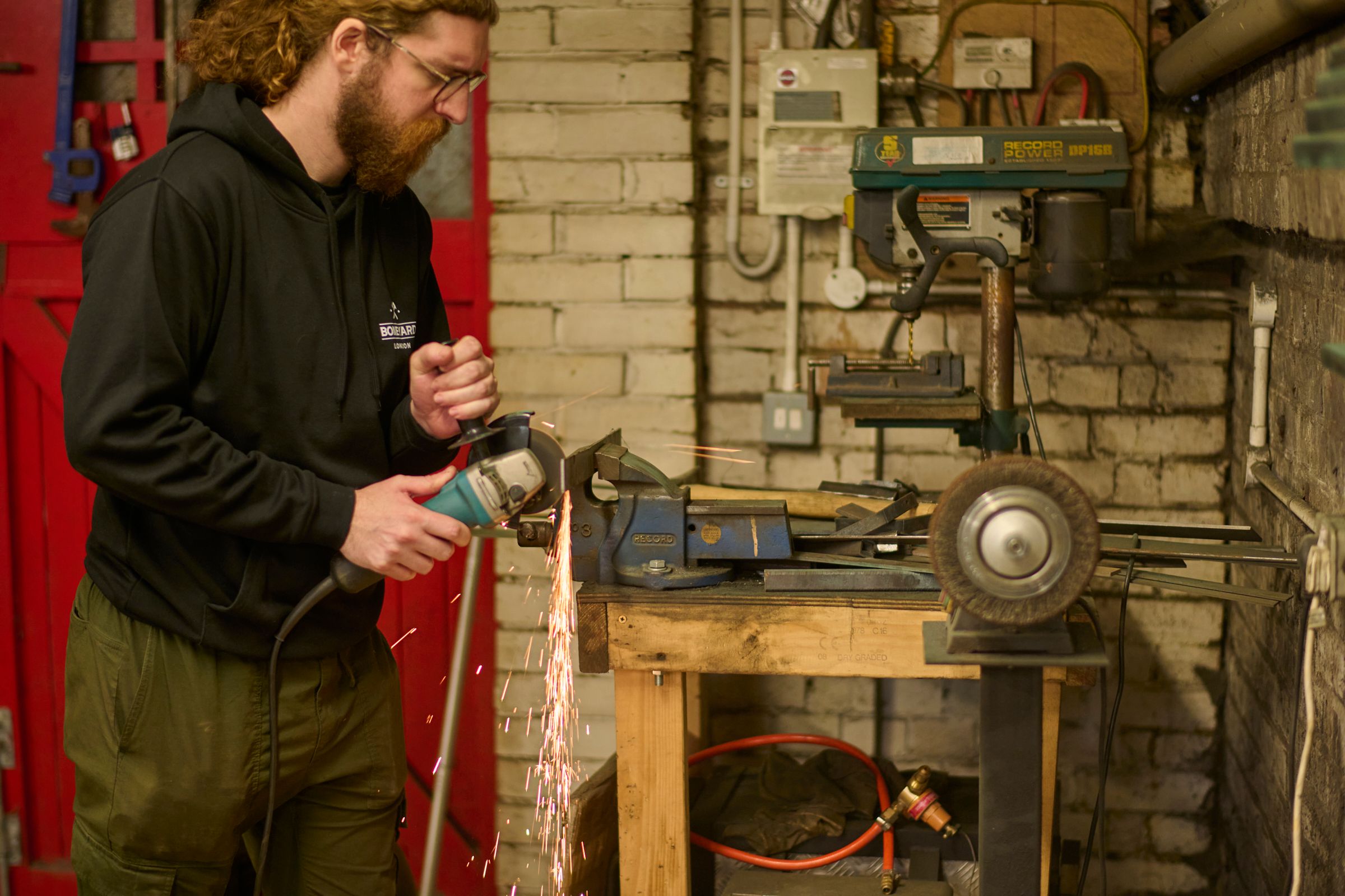 A bearded man wearing glasses uses an angle grinder, producing sparks, in a workshop.