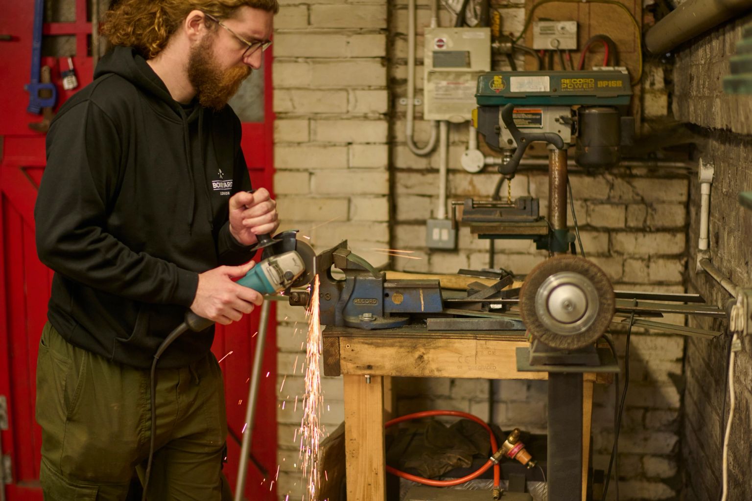 A man with a beard and glasses uses an angle grinder, creating sparks, in a workshop.