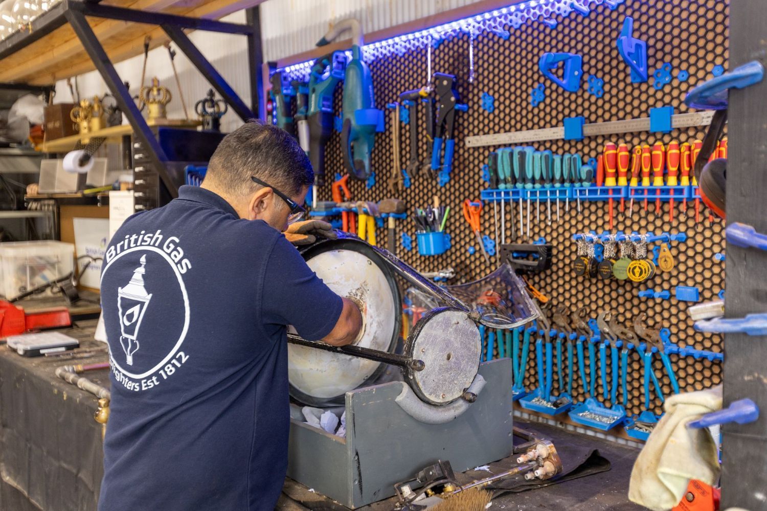 A man in a British Gas Lighters shirt works on a circular metal object in a workshop filled with tools.