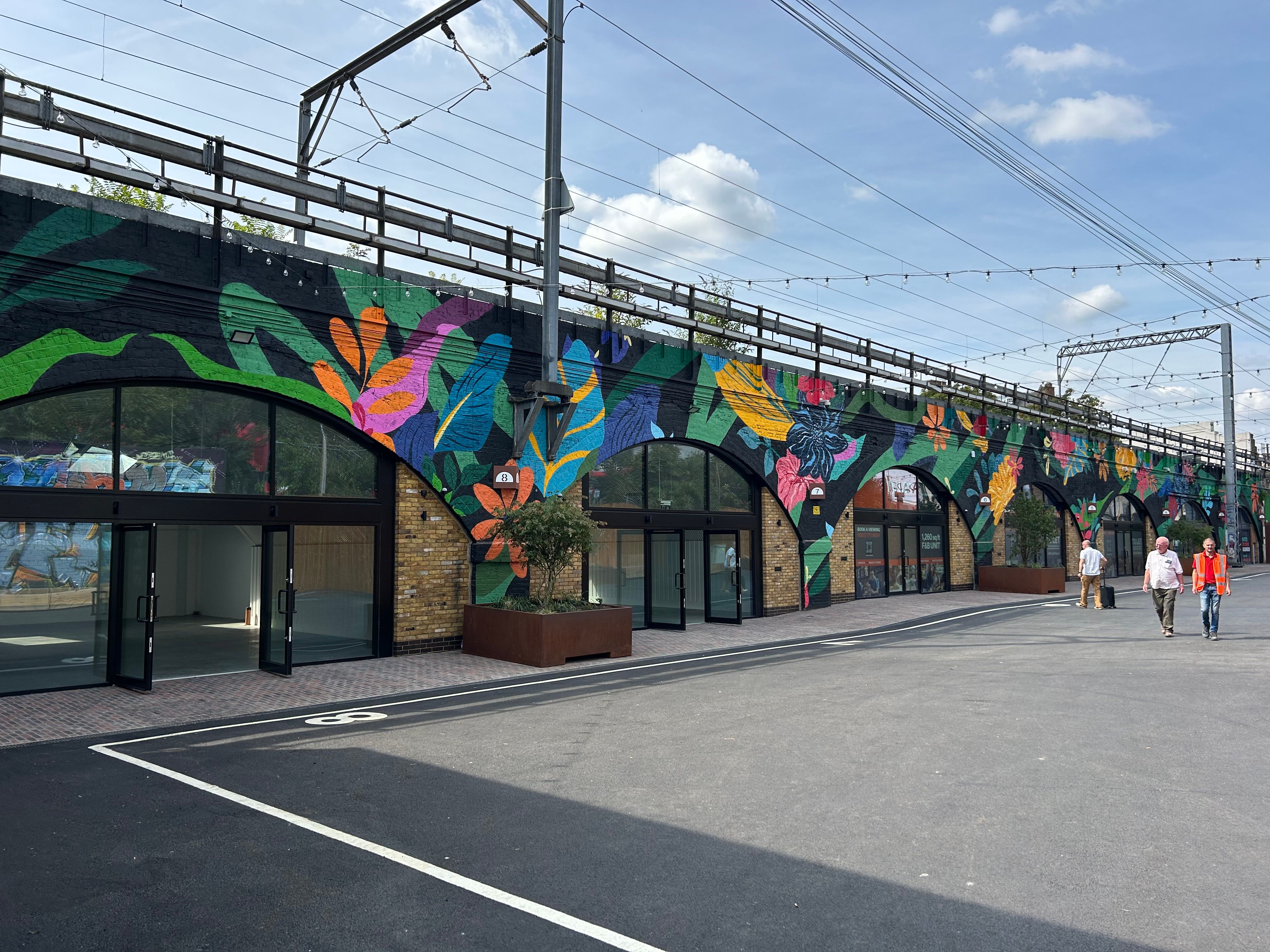 A long railway viaduct with a vibrant tropical mural on its arched walls above several empty shopfronts, with a paved area and people walking.