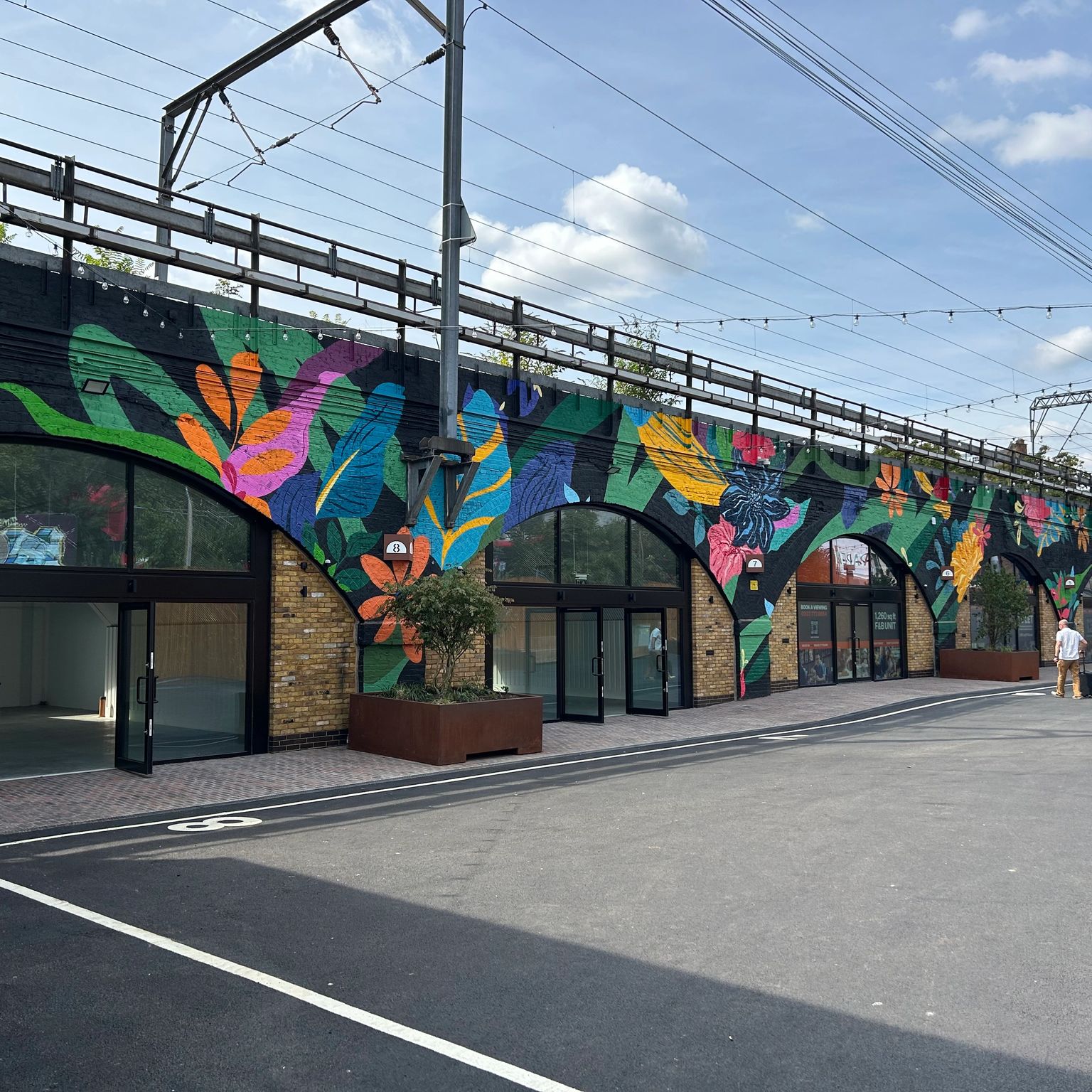 A long railway viaduct with a vibrant tropical mural on its arched walls above several empty shopfronts, with a paved area and people walking.